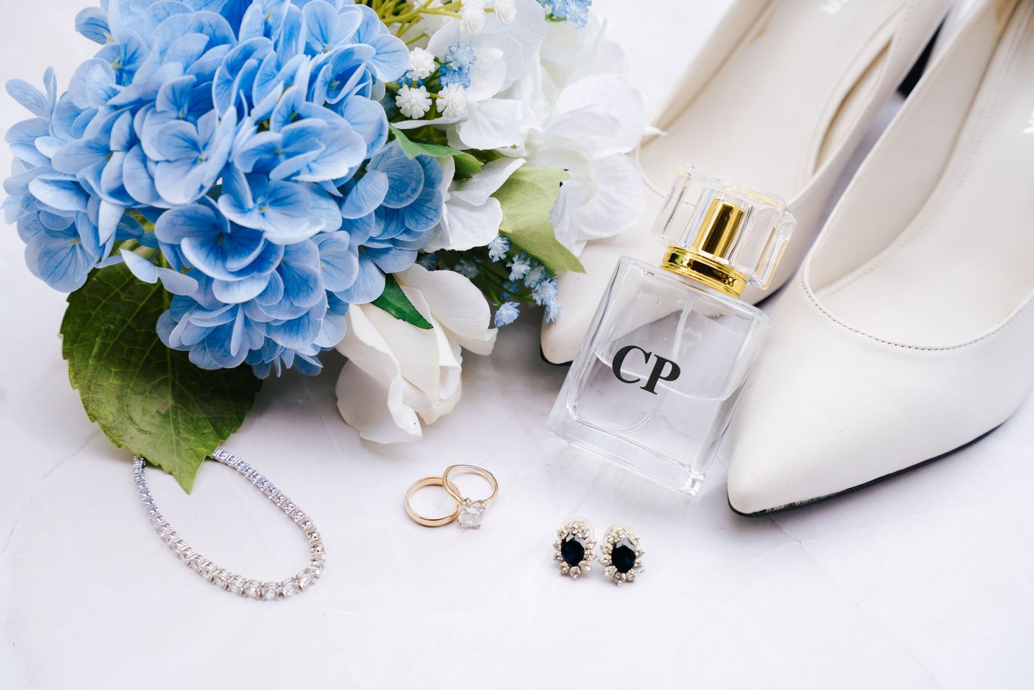 Wedding accessories including blue hydrangeas, white heels, perfume bottle, pearl necklace, and wedding rings on marble surface.
