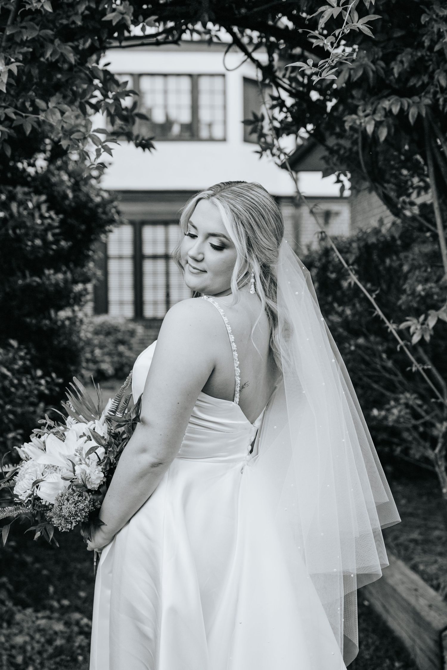 A bride in a flowing white gown poses with her bouquet against a garden backdrop.