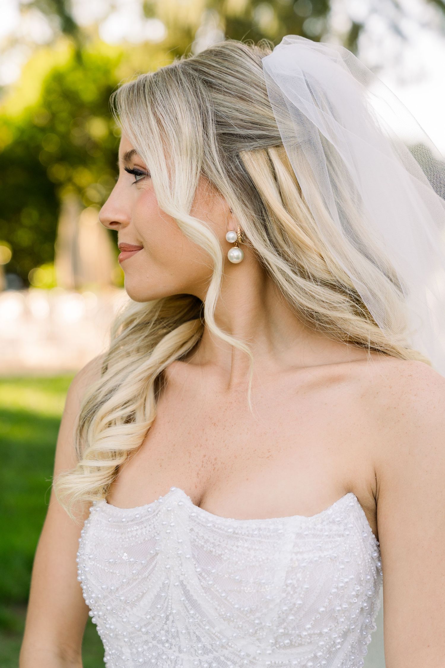 Blonde bride in strapless pearl wedding gown with veil and pearl drop earrings, smiling outdoors.