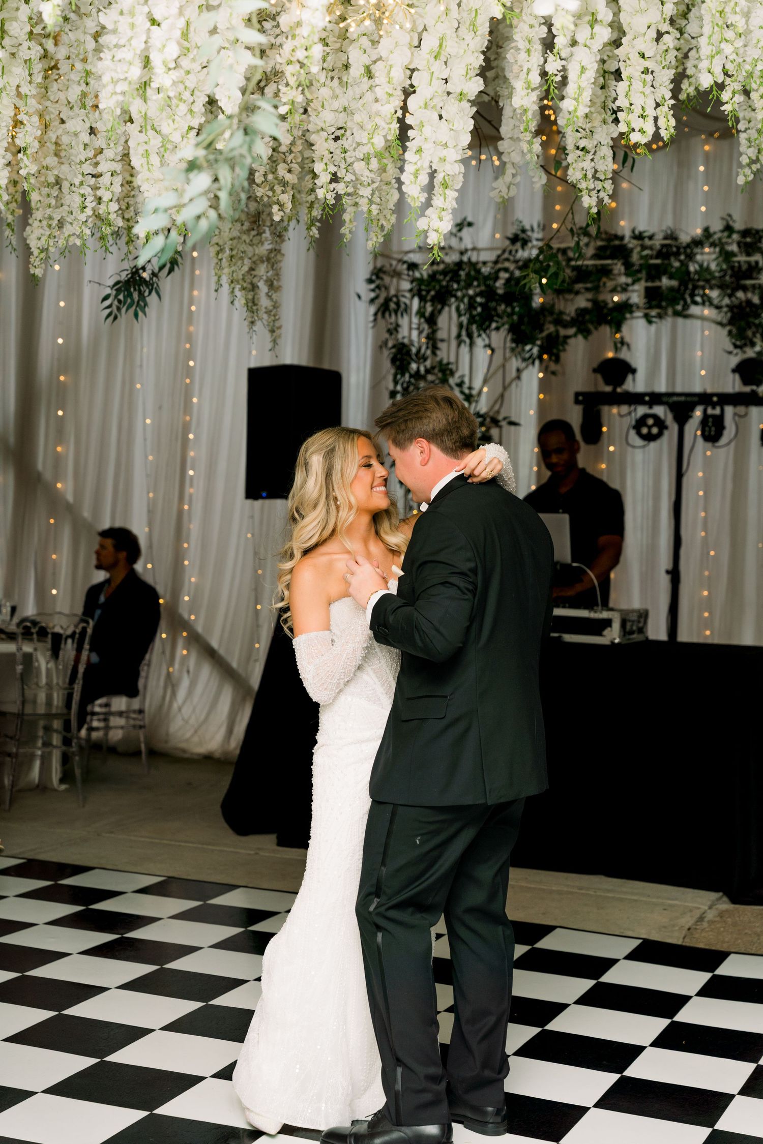 Bride and groom share first dance on black and white checkered floor under white floral ceiling decorations.