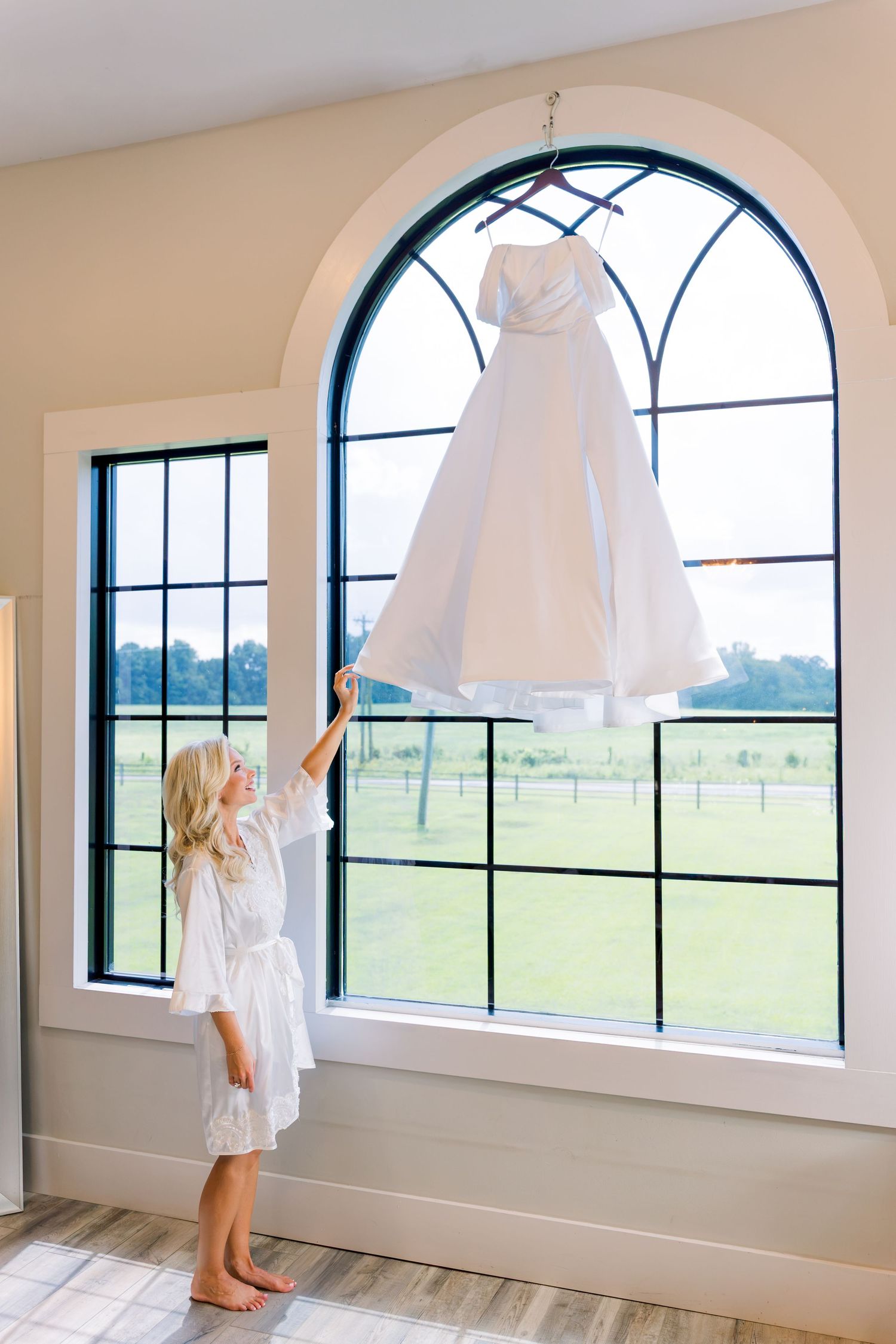 Young flower girl in white robe reaching up toward a wedding dress hanging in an arched window.