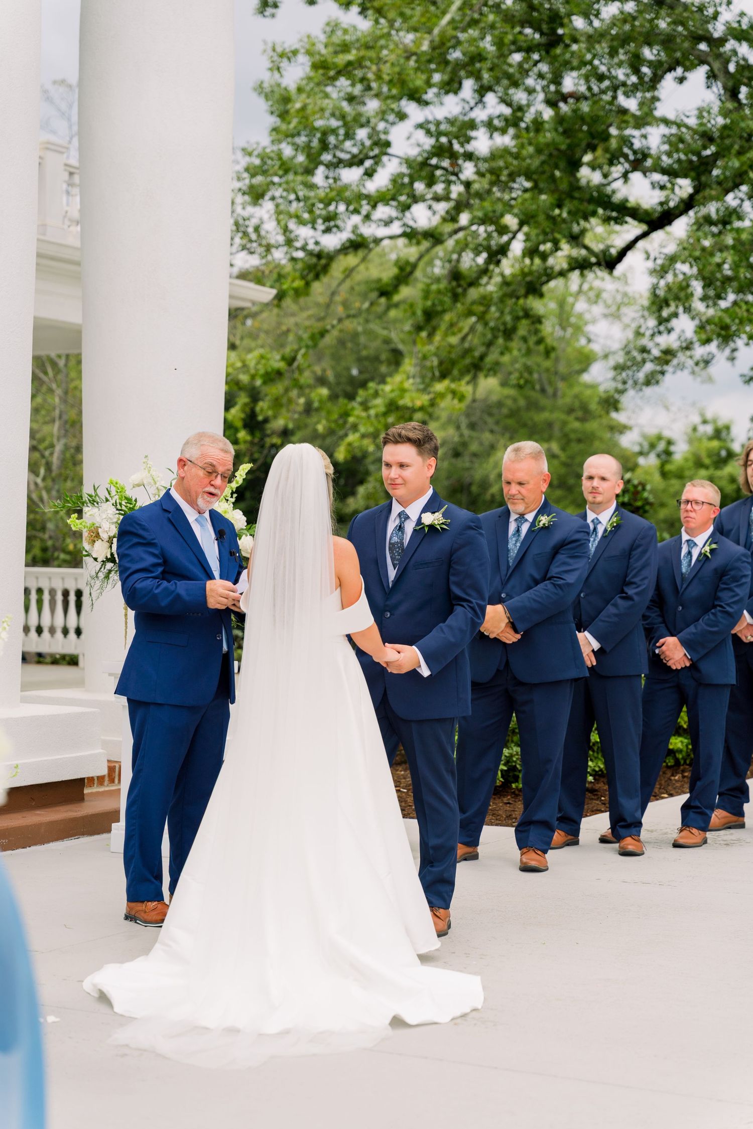 Outdoor wedding ceremony with bride and groom holding hands before officiant, groomsmen in navy suits standing beside white columns.