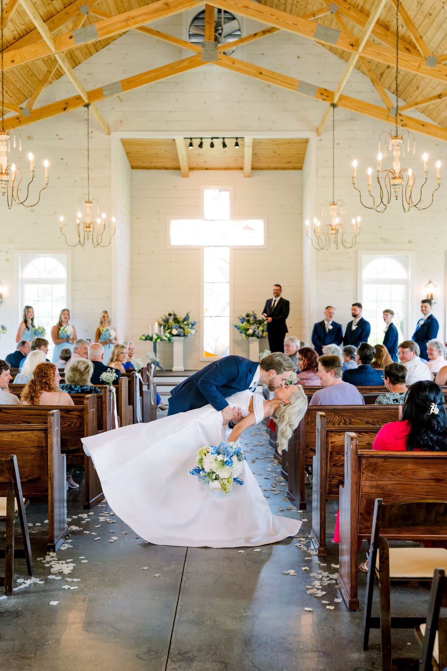 Bride and groom share a romantic dip kiss in a white chapel aisle, surrounded by seated wedding guests and a glowing cross.