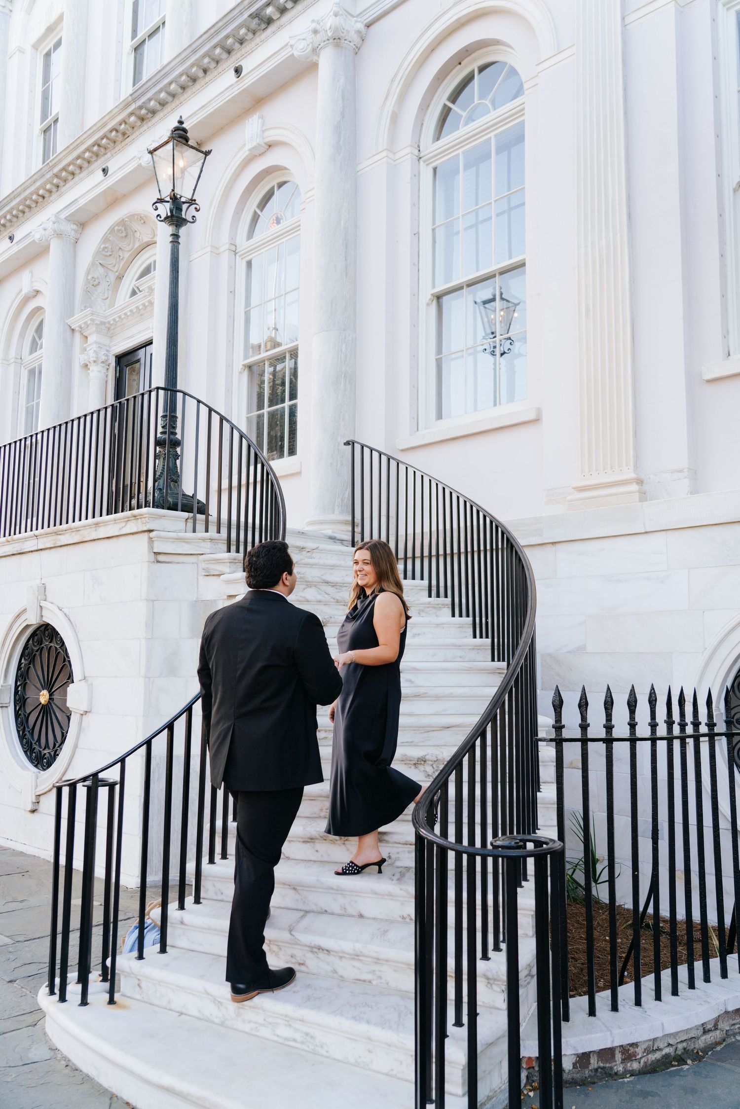 Couple poses on white marble steps with black iron railings in front of a historic building.