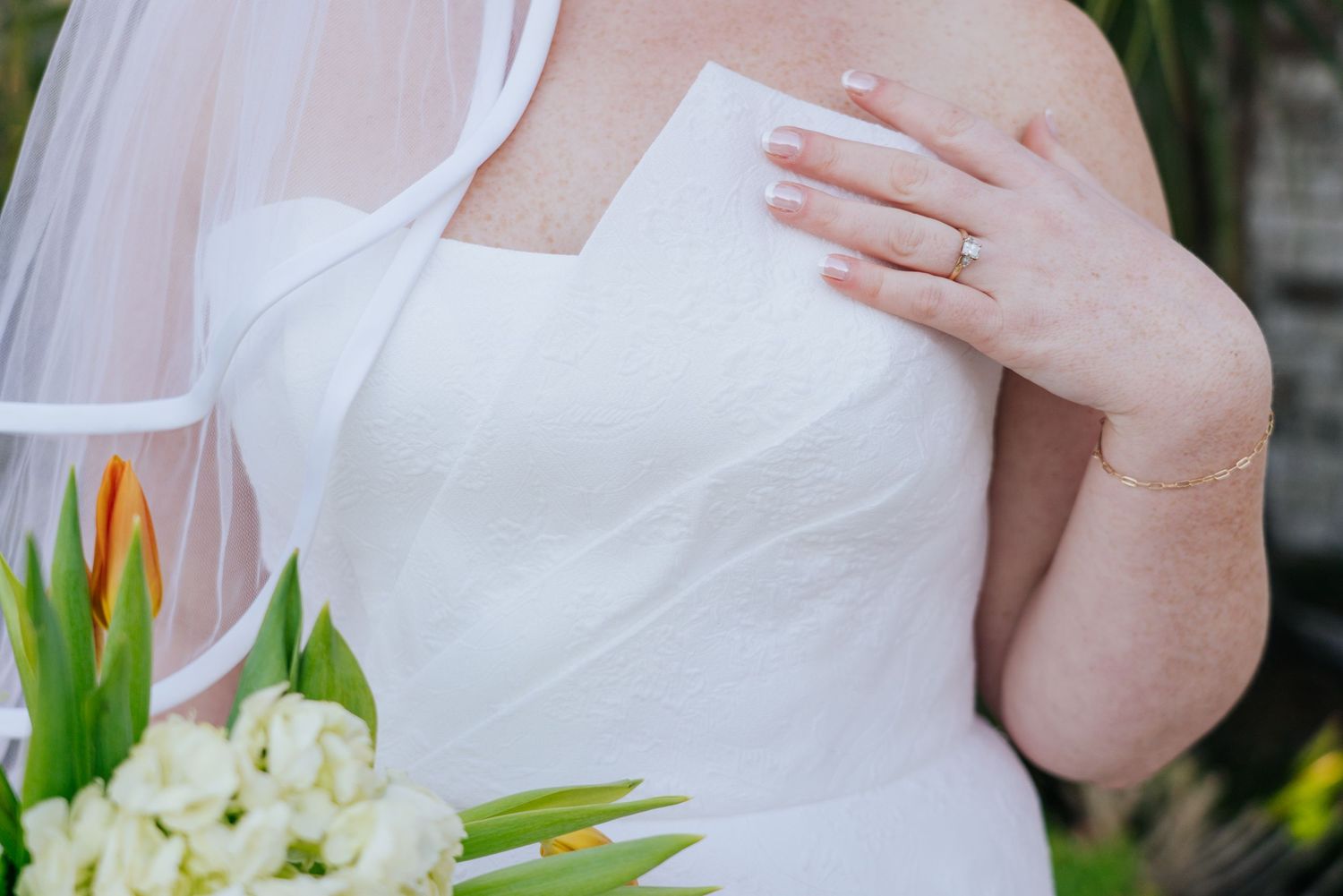 A white bridal gown and bouquet are shown in an elegant outdoor setting with brick walls and palm trees.