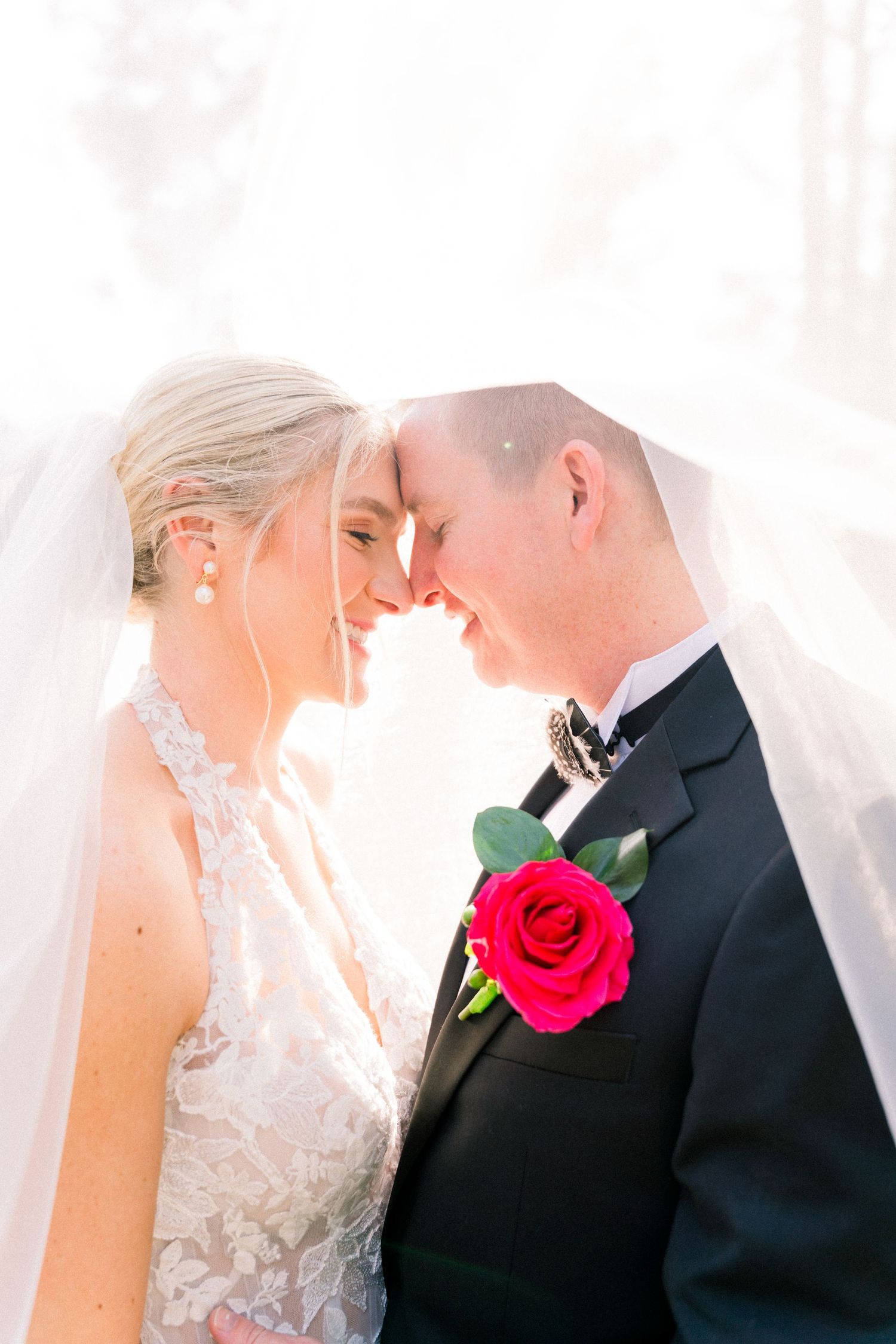 Bride and groom touching foreheads under veil, groom wearing black tuxedo with red rose boutonniere.