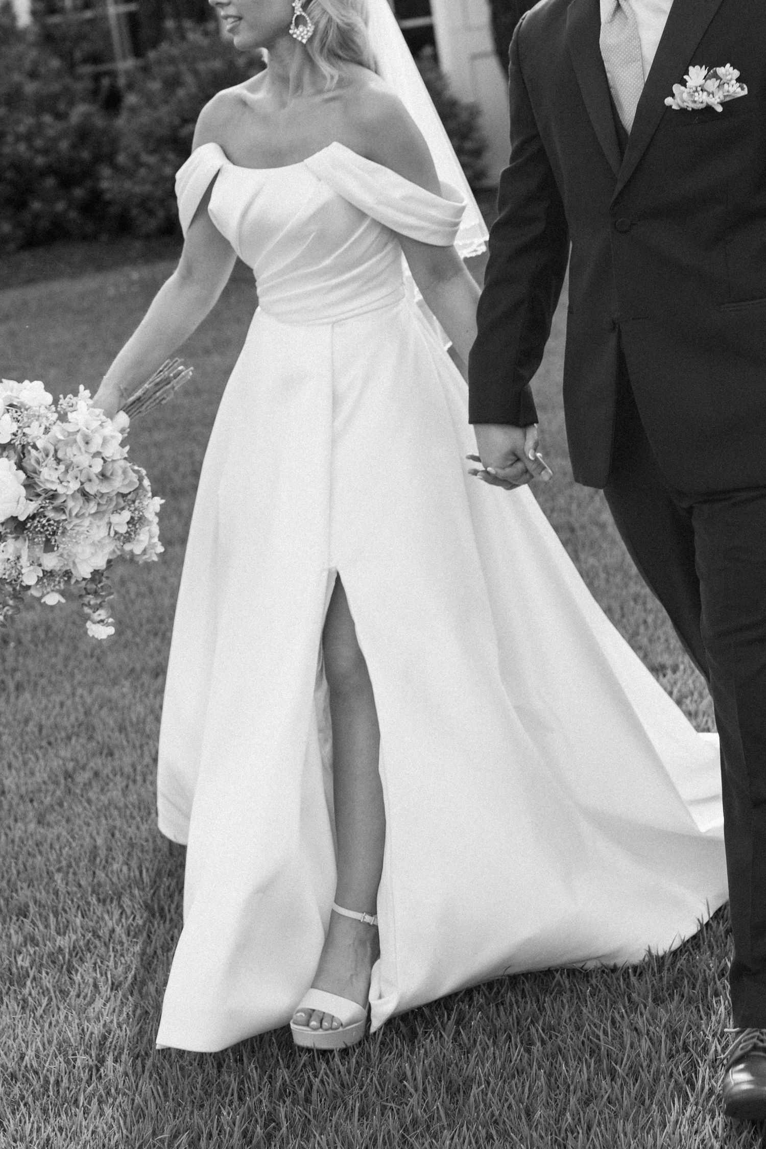 Bride in off-shoulder white gown with front slit holds bouquet while walking with groom in black suit.