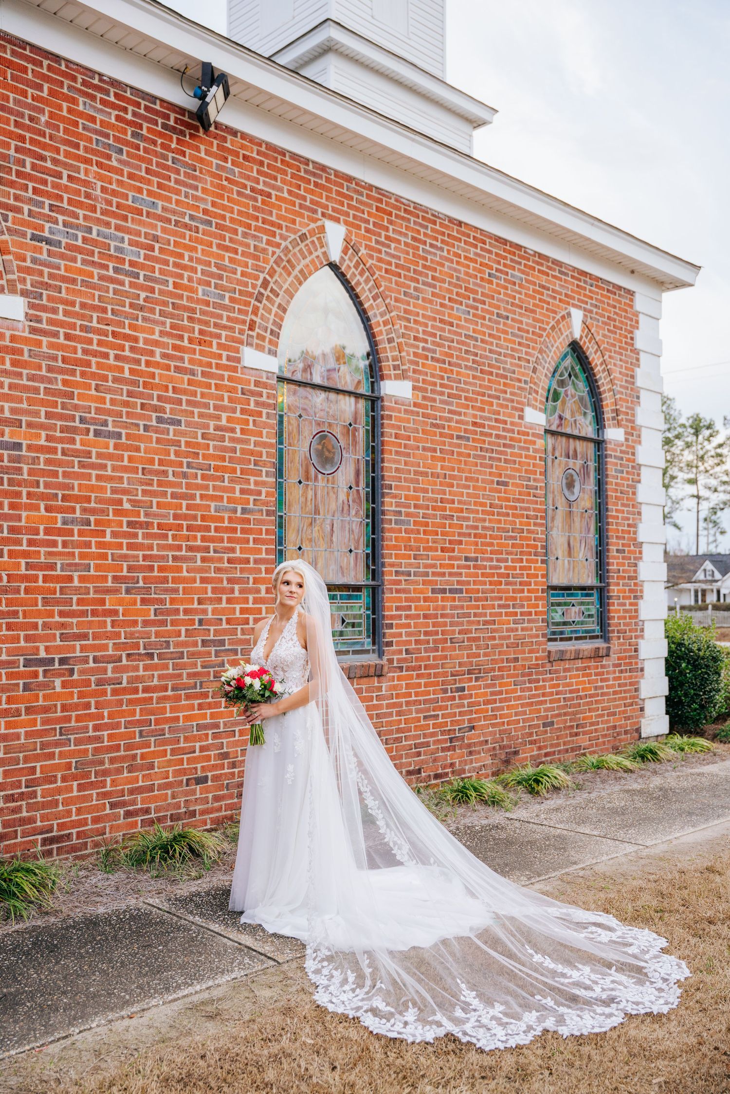 A bride in a flowing white dress and veil poses elegantly against a red brick church wall with arched windows.