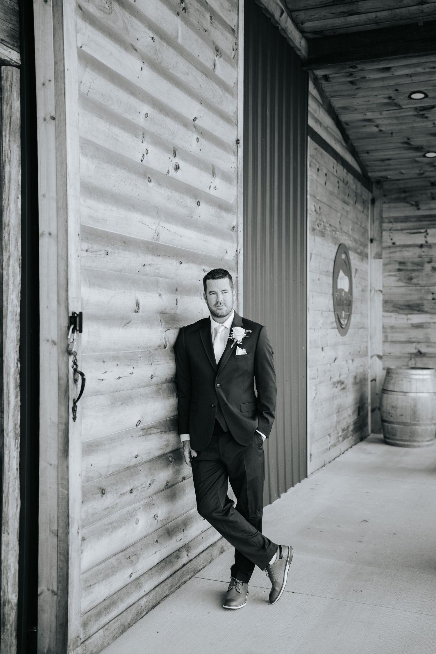 Groom in formal black suit poses in modern rustic hallway.
