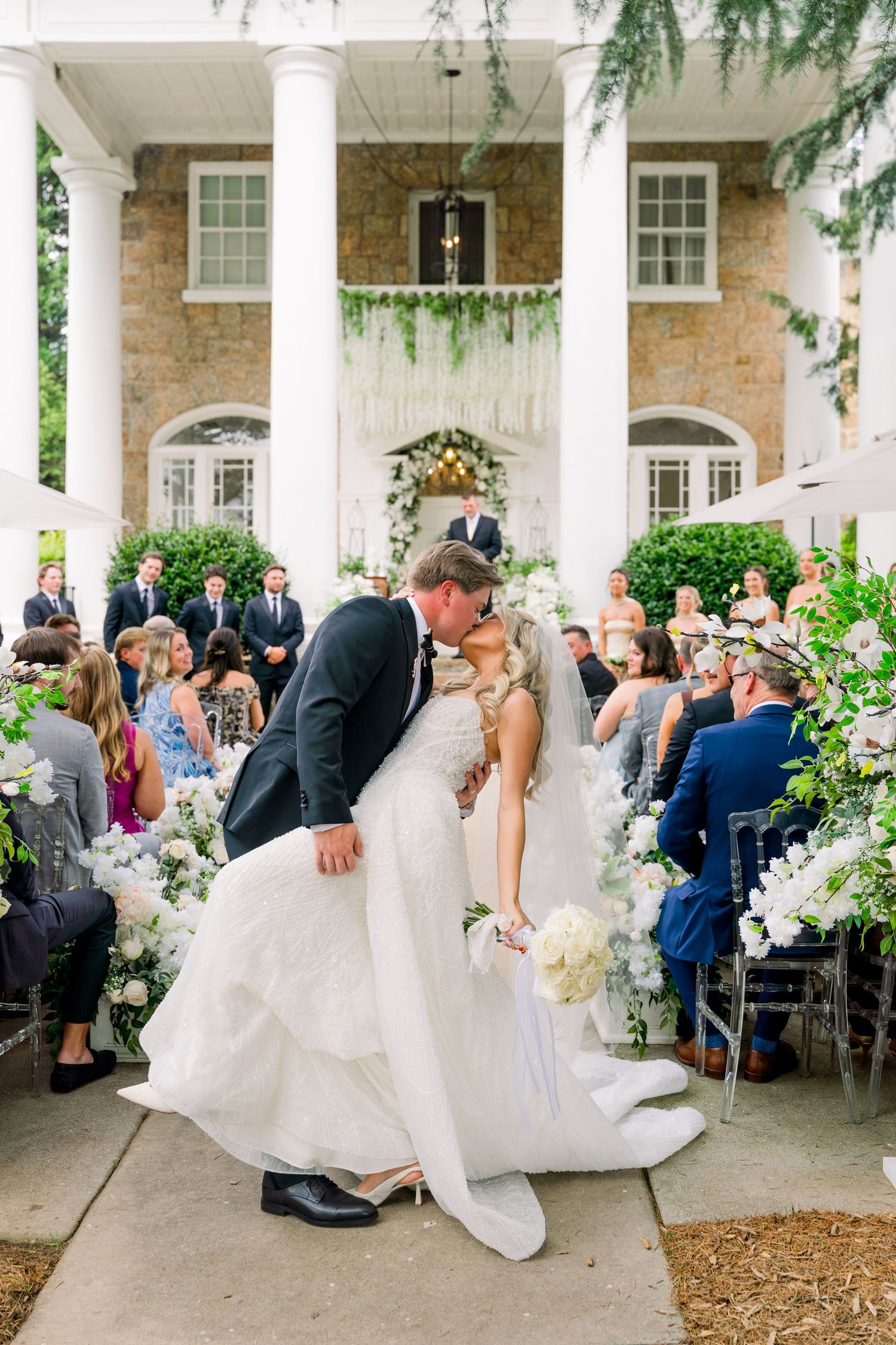 Bride and groom share first kiss at outdoor wedding ceremony with white floral arrangements and colonial venue backdrop.