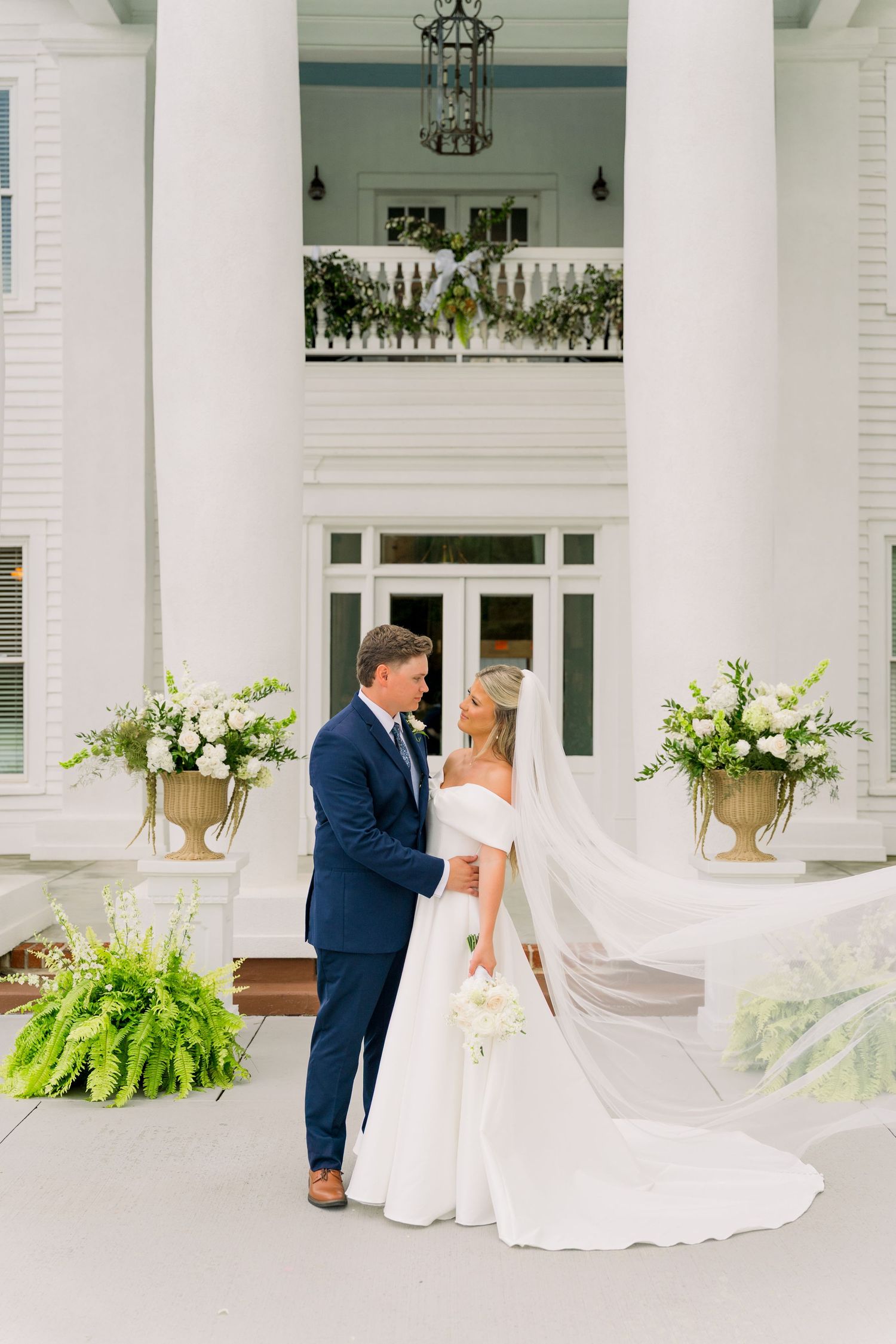 Bride and groom share a romantic moment outside a white colonial venue with floral urns and a flowing veil.