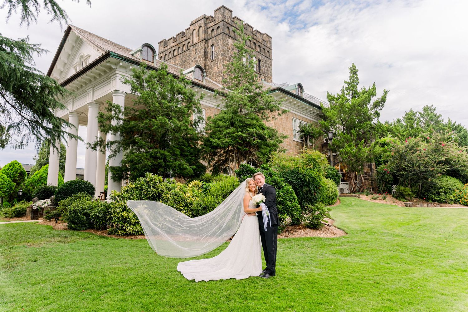 Bride and groom kissing on lawn with veil flowing, elegant stone mansion venue backdrop on wedding day.