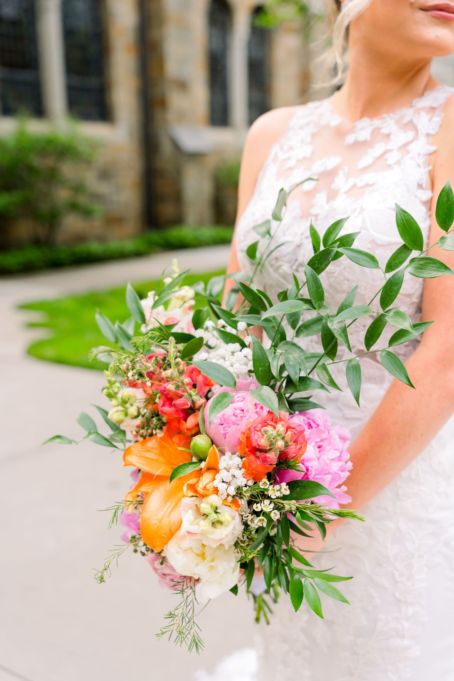 Bride holding vibrant bridal bouquet with pink peonies, orange lilies, and lush greenery in lace wedding gown.
