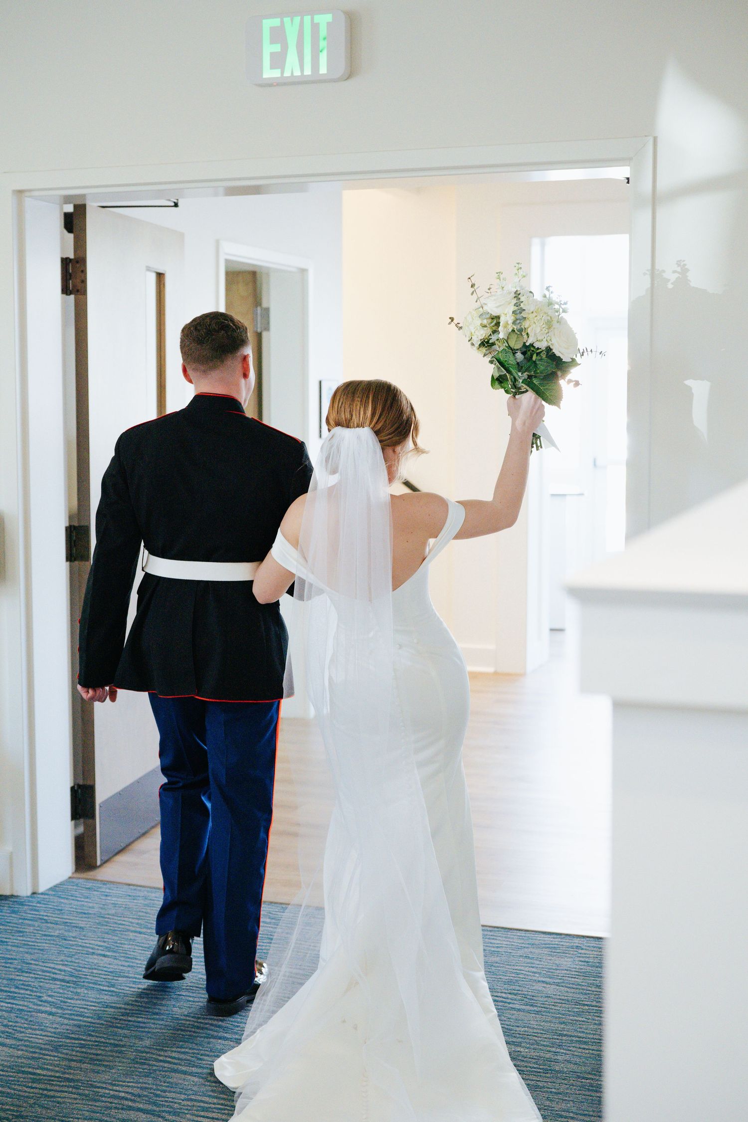 Wedding couple walking down a bright hallway with an exit sign.