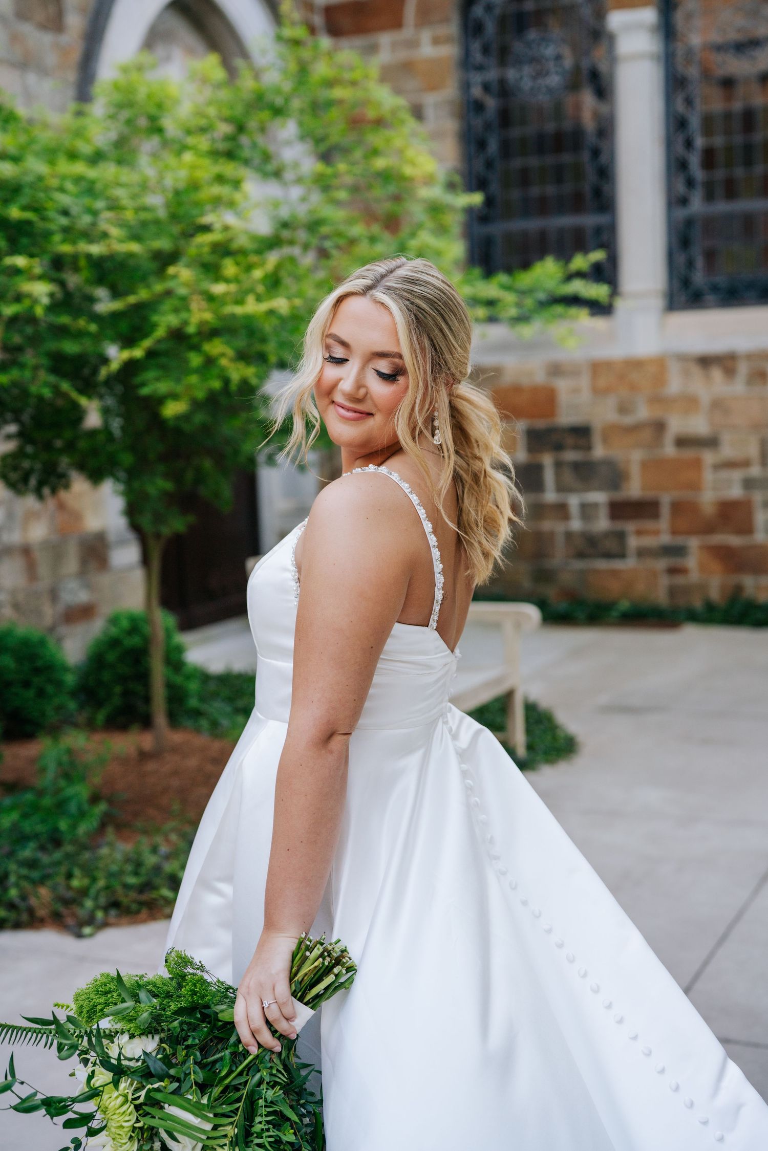 Elegant white wedding dress photographed in front of a stone church with lush greenery.