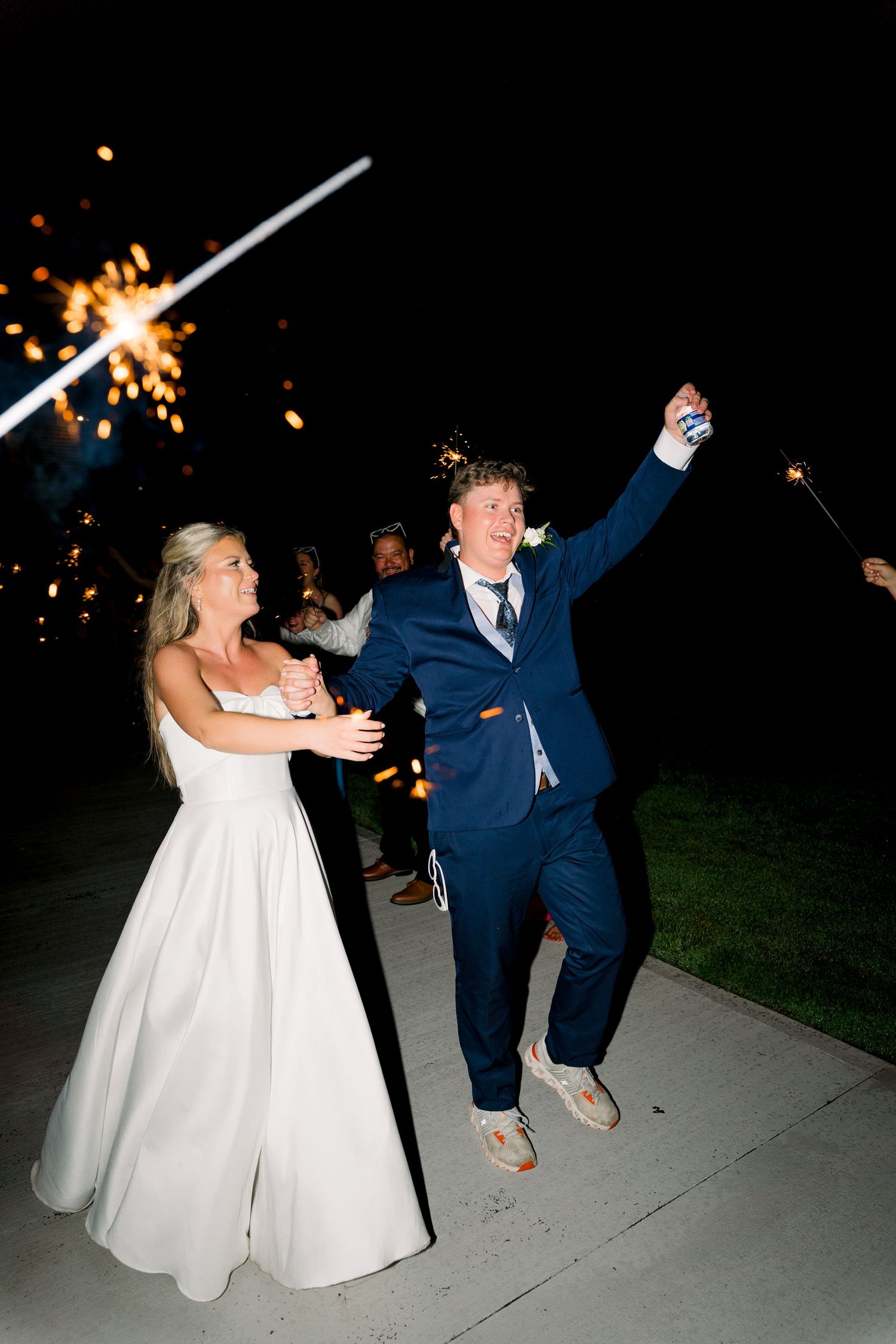 Joyful newlyweds exit wedding reception through sparkler tunnel, bride in white gown, groom in navy suit, night celebration.