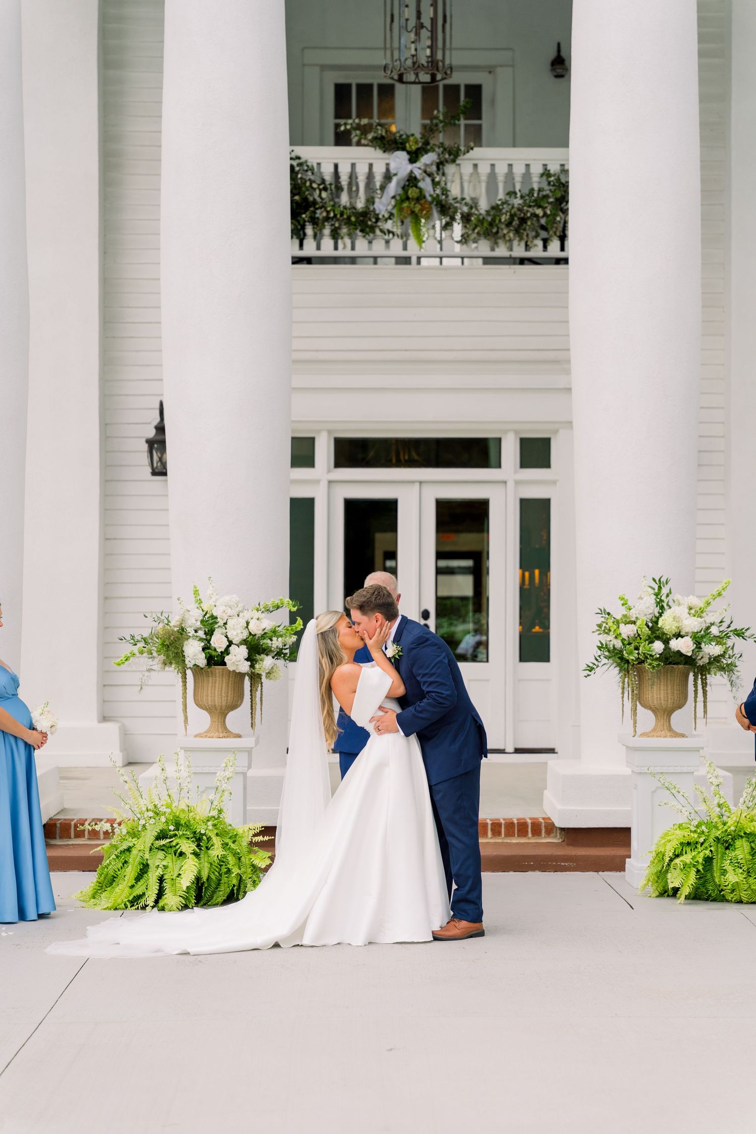 Bride and groom share first kiss at elegant white colonial venue with floral arrangements and tall columns.
