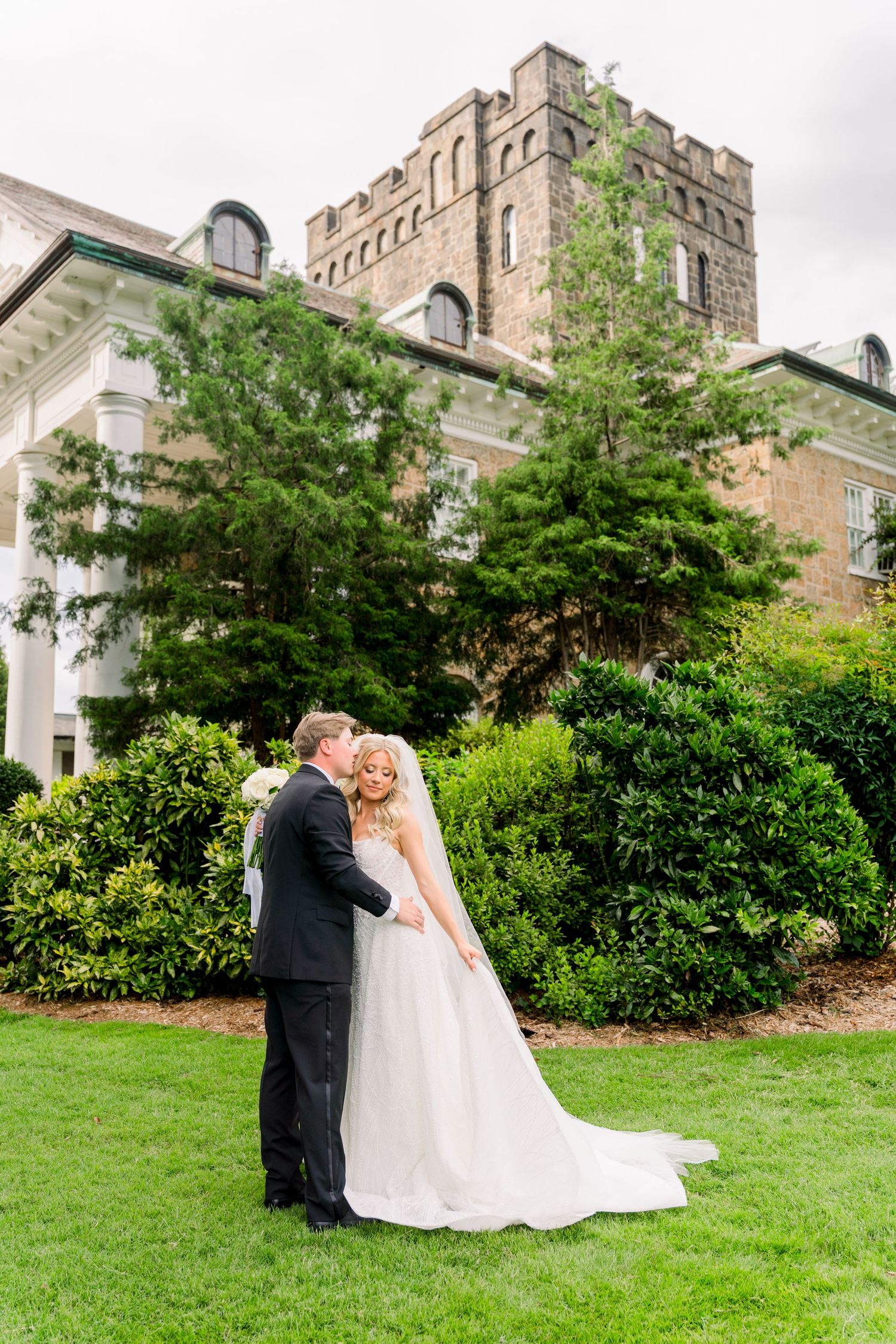 A bride and groom share a romantic embrace on a lush green lawn in front of an elegant stone castle venue.