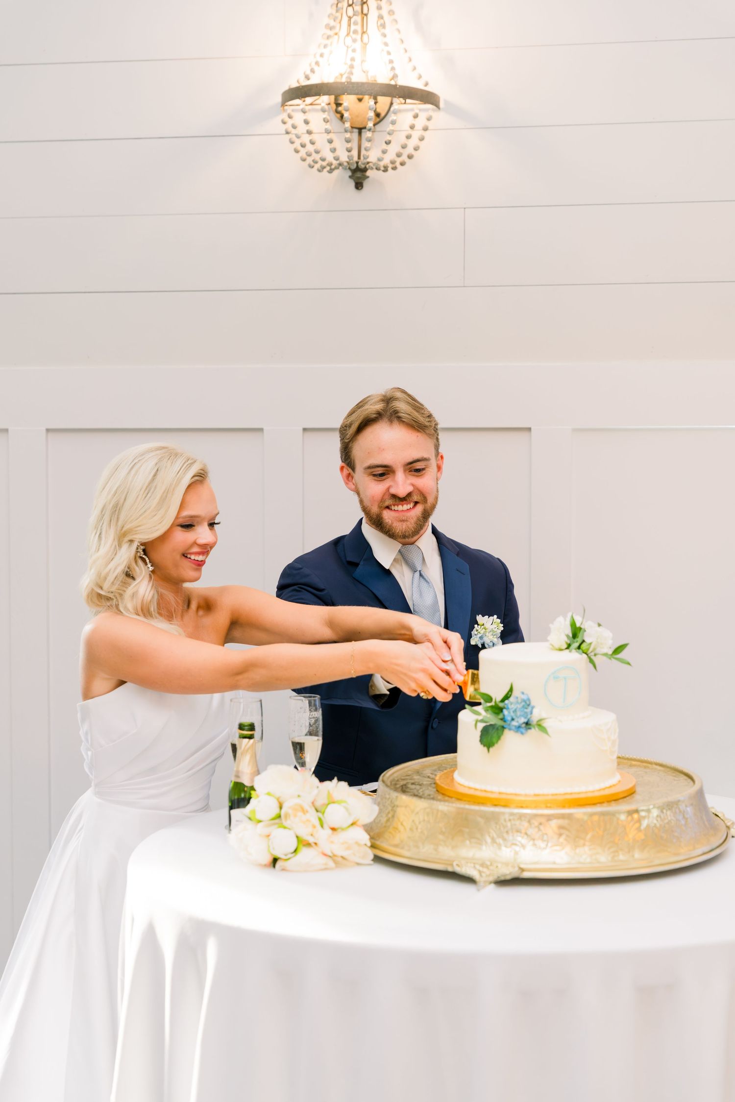 Bride and groom smiling while cutting their elegant two-tier white wedding cake adorned with blue flowers.