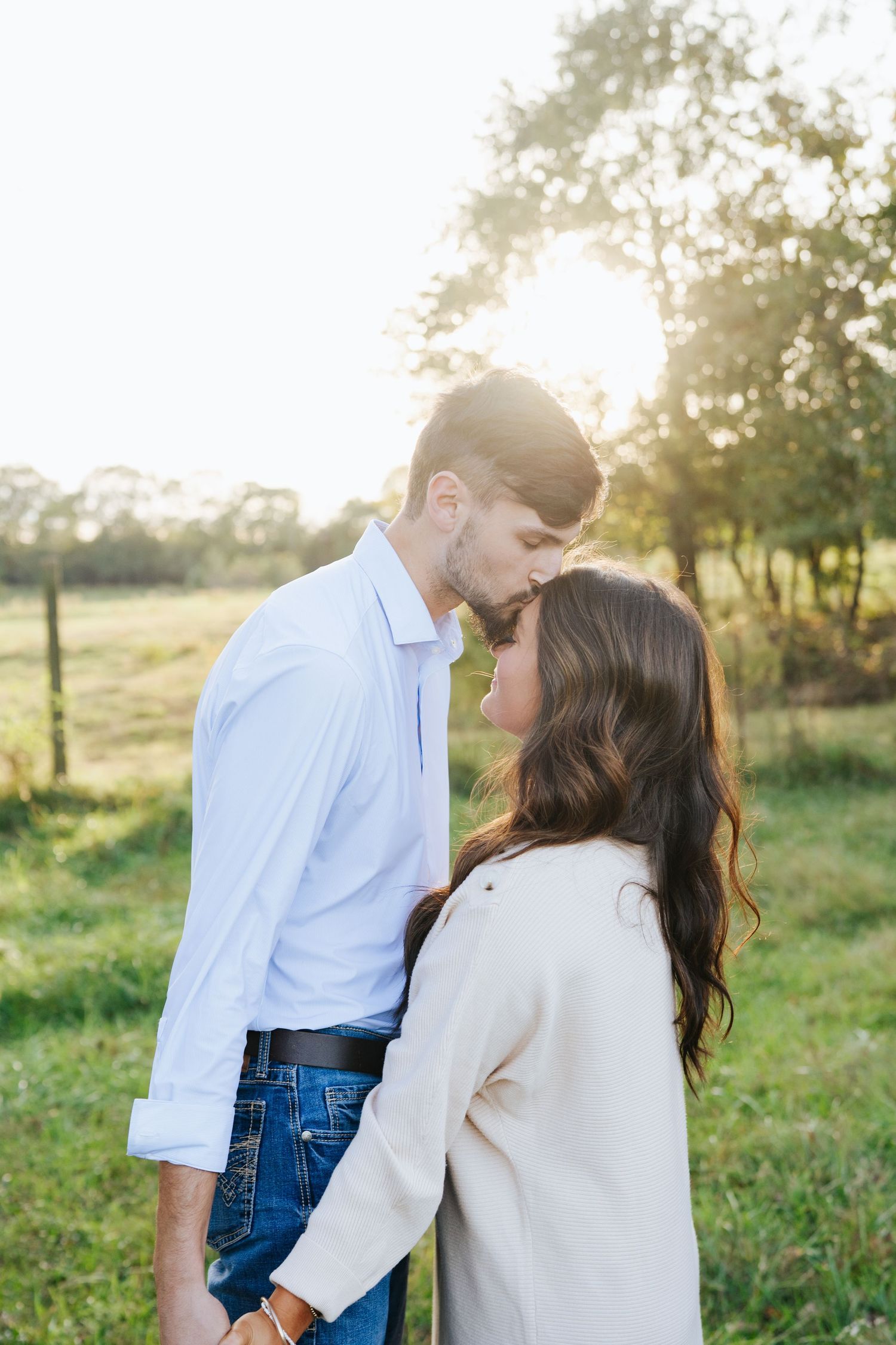 A romantic couple embraces in a sunlit meadow during golden hour while wearing casual white and blue outfits.