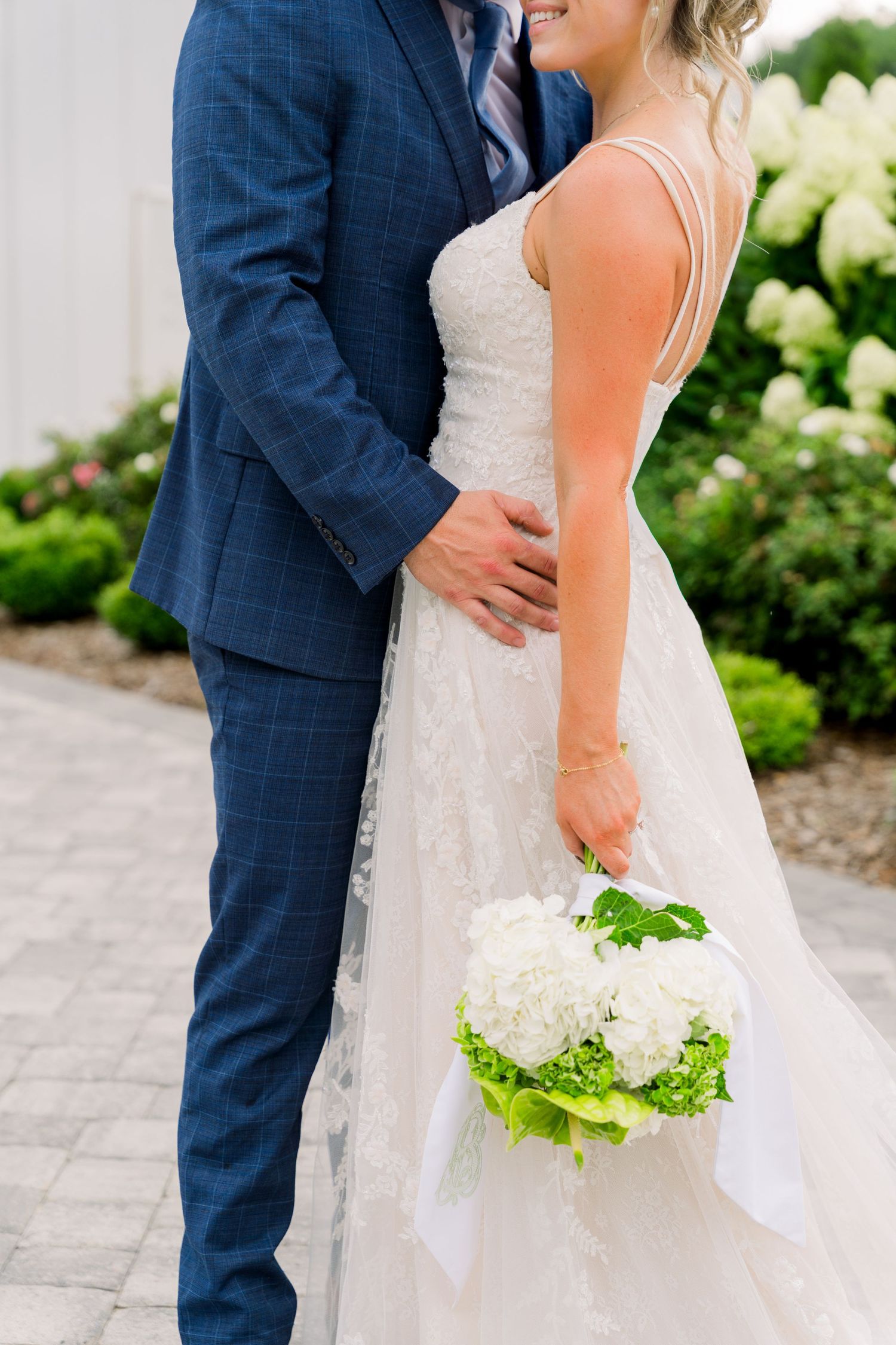 Bride in white lace gown holding white hydrangea bouquet embraced by groom in blue plaid suit outdoors.