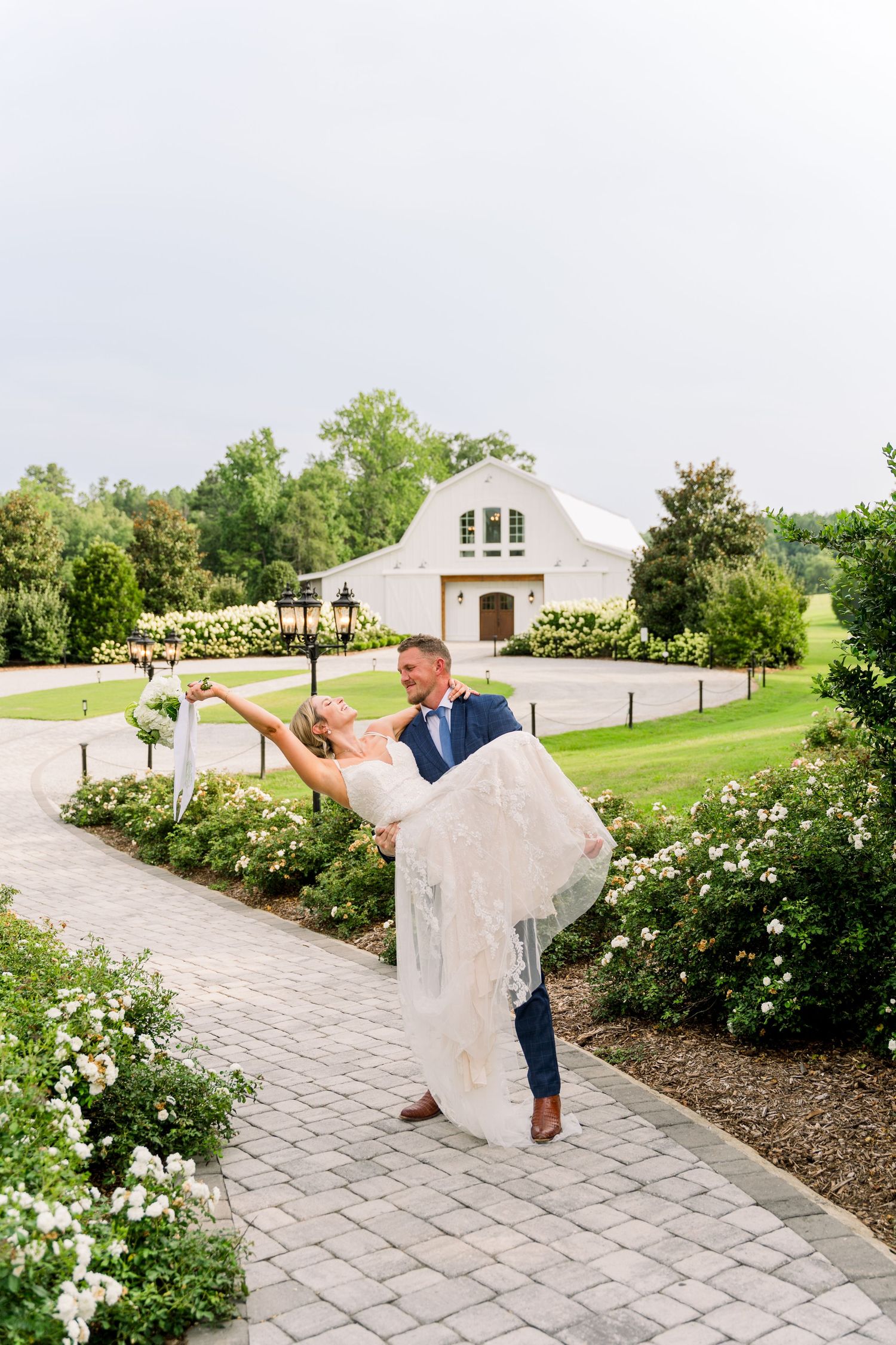 Groom lifting bride on a brick path at a white barn wedding venue surrounded by lush greenery and white flowers.