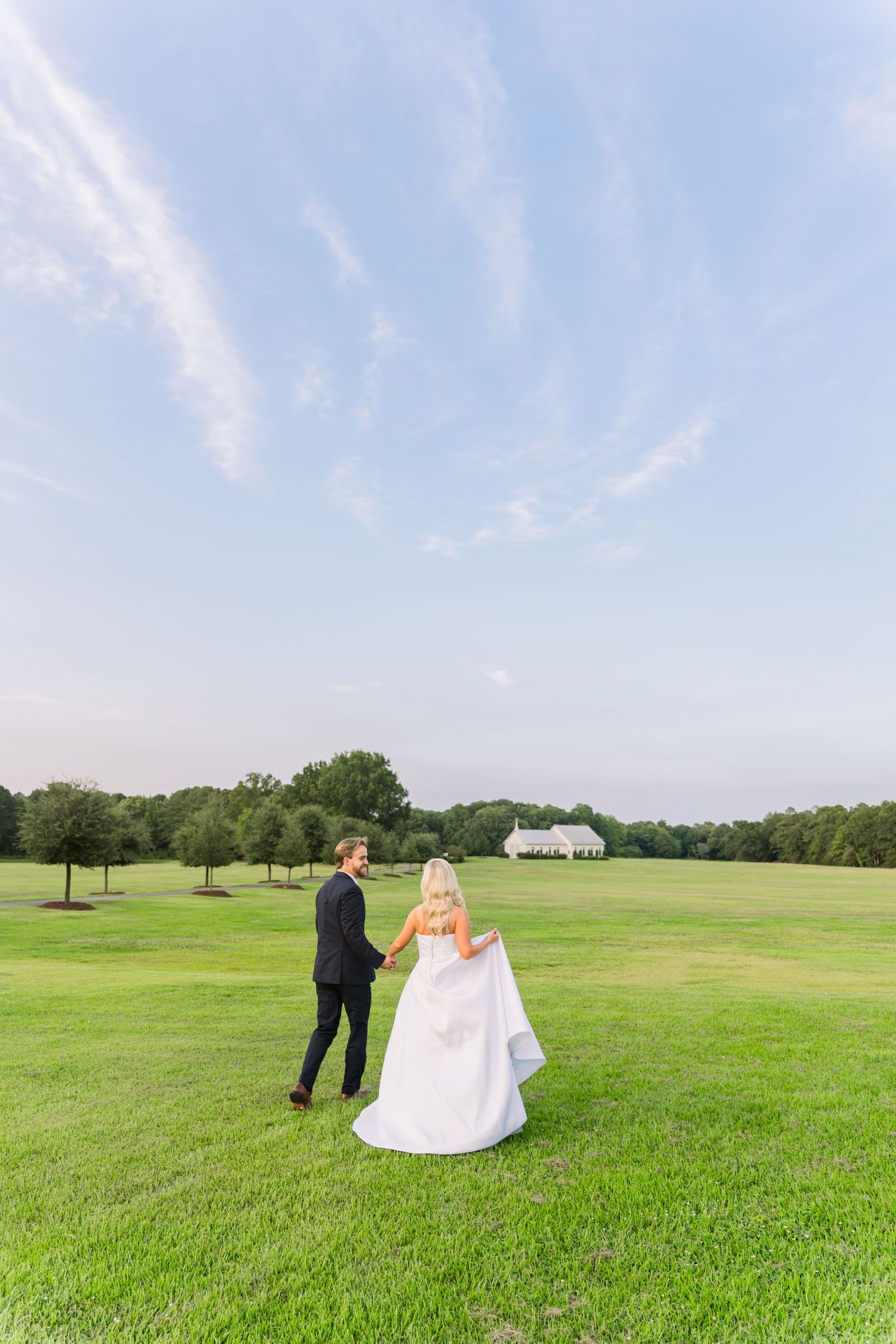 Bride and groom walking hand in hand across a lush green field with a white venue in the distance.