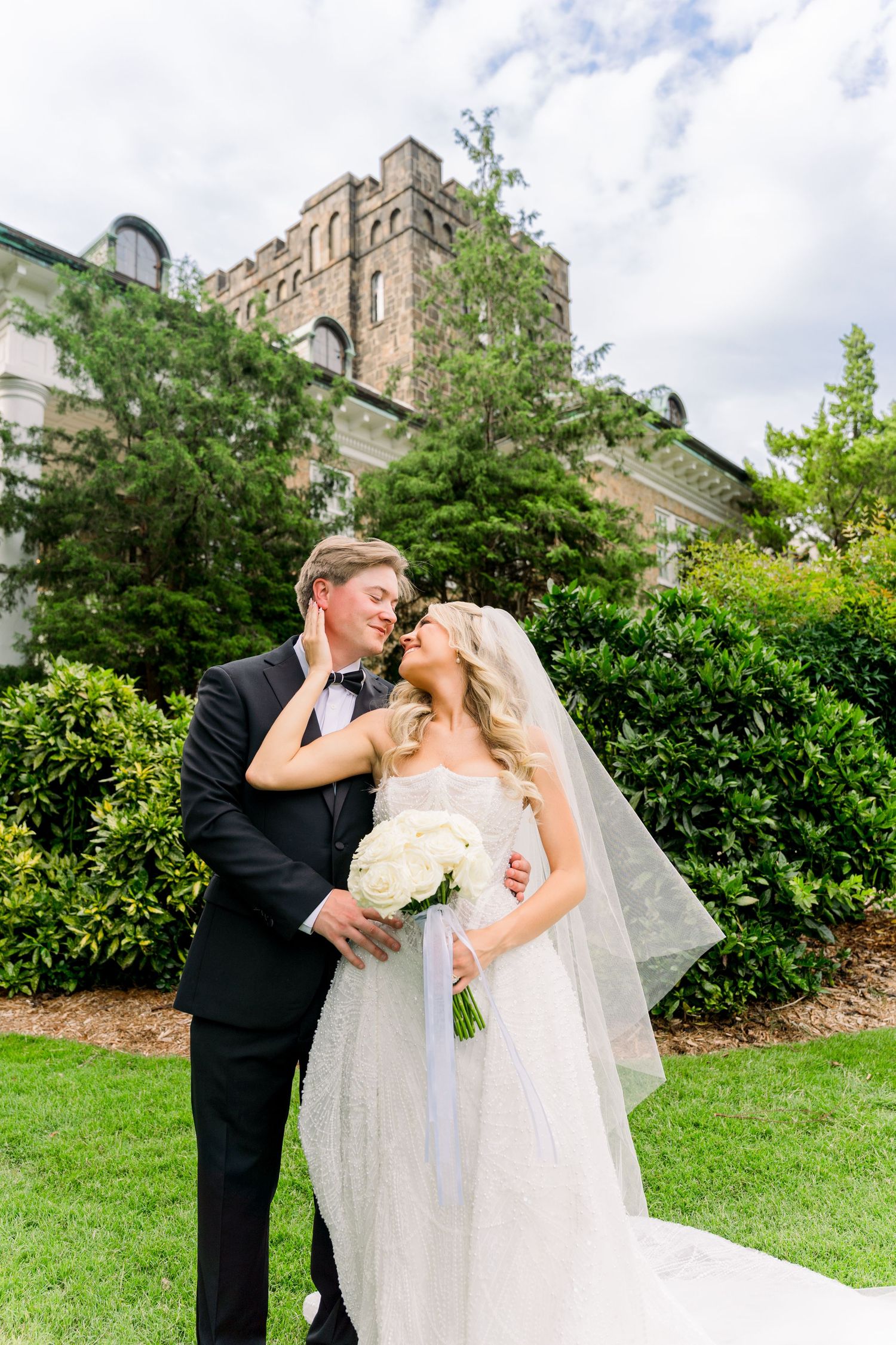 Bride and groom share a romantic moment in garden, bride holding white bouquet in strapless gown with veil, groom in black tux.