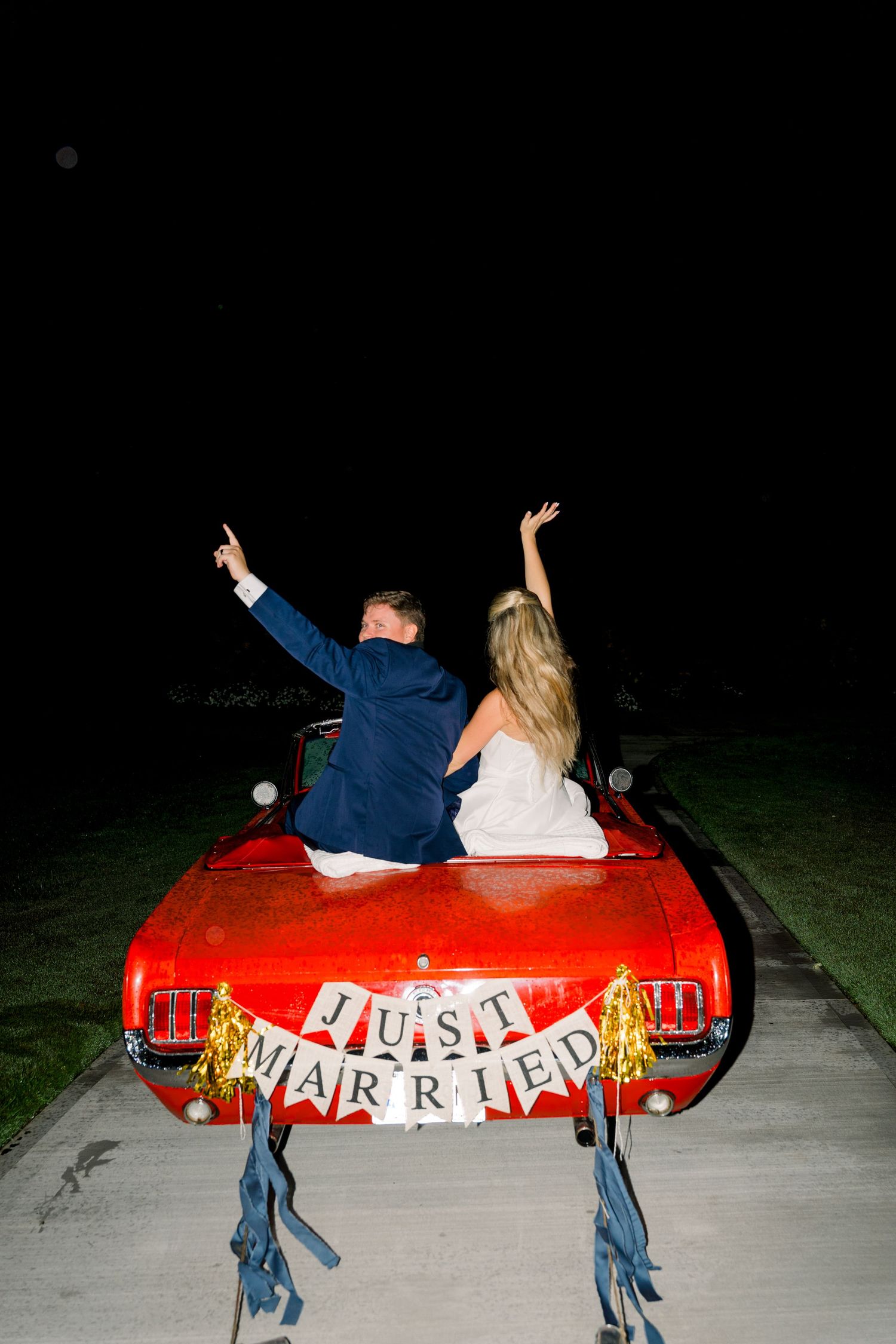 Newlyweds celebrate in a red convertible with a Just Married sign at night, raising their hands in joy.