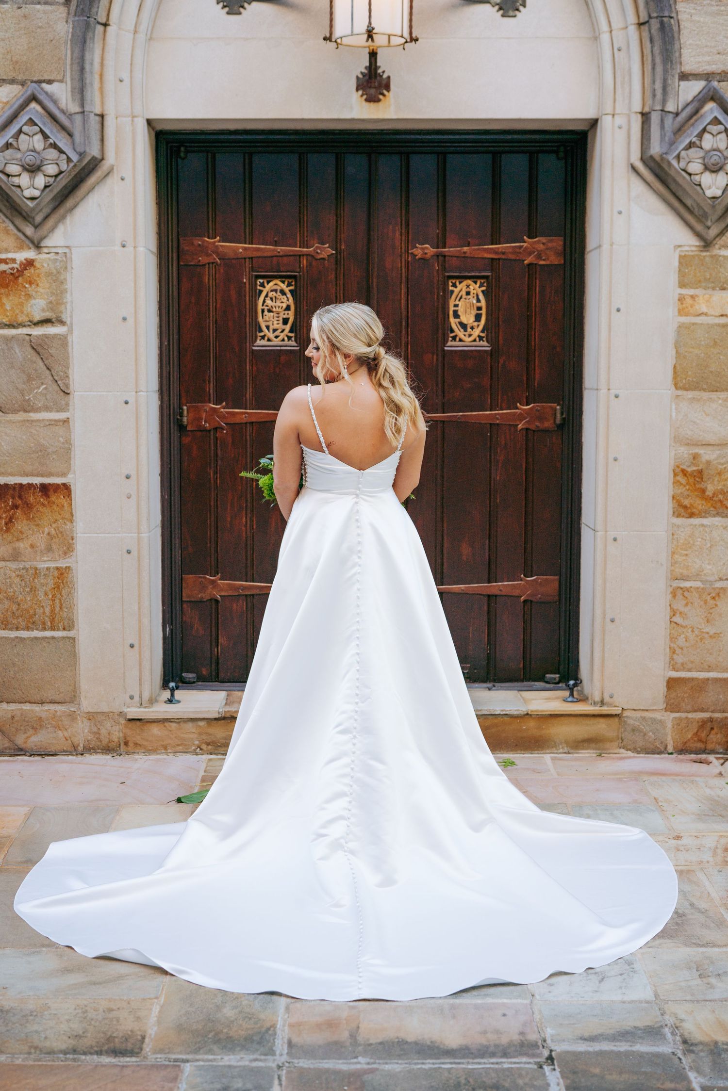 Back view of flowing white wedding dress with long train against historic stone church doors.