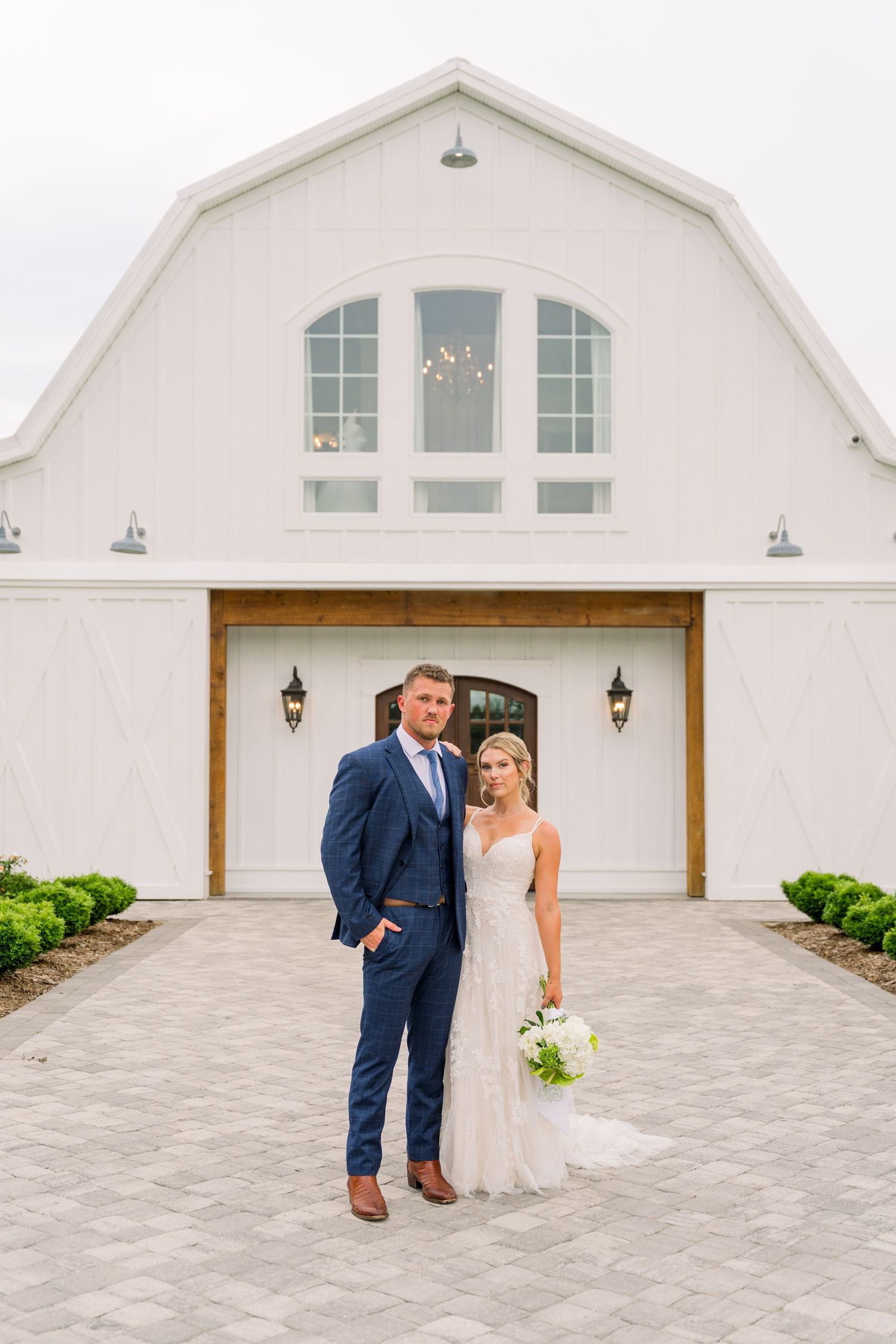Bride and groom posing in front of a white barn wedding venue, bride holding white bouquet, groom in navy suit.