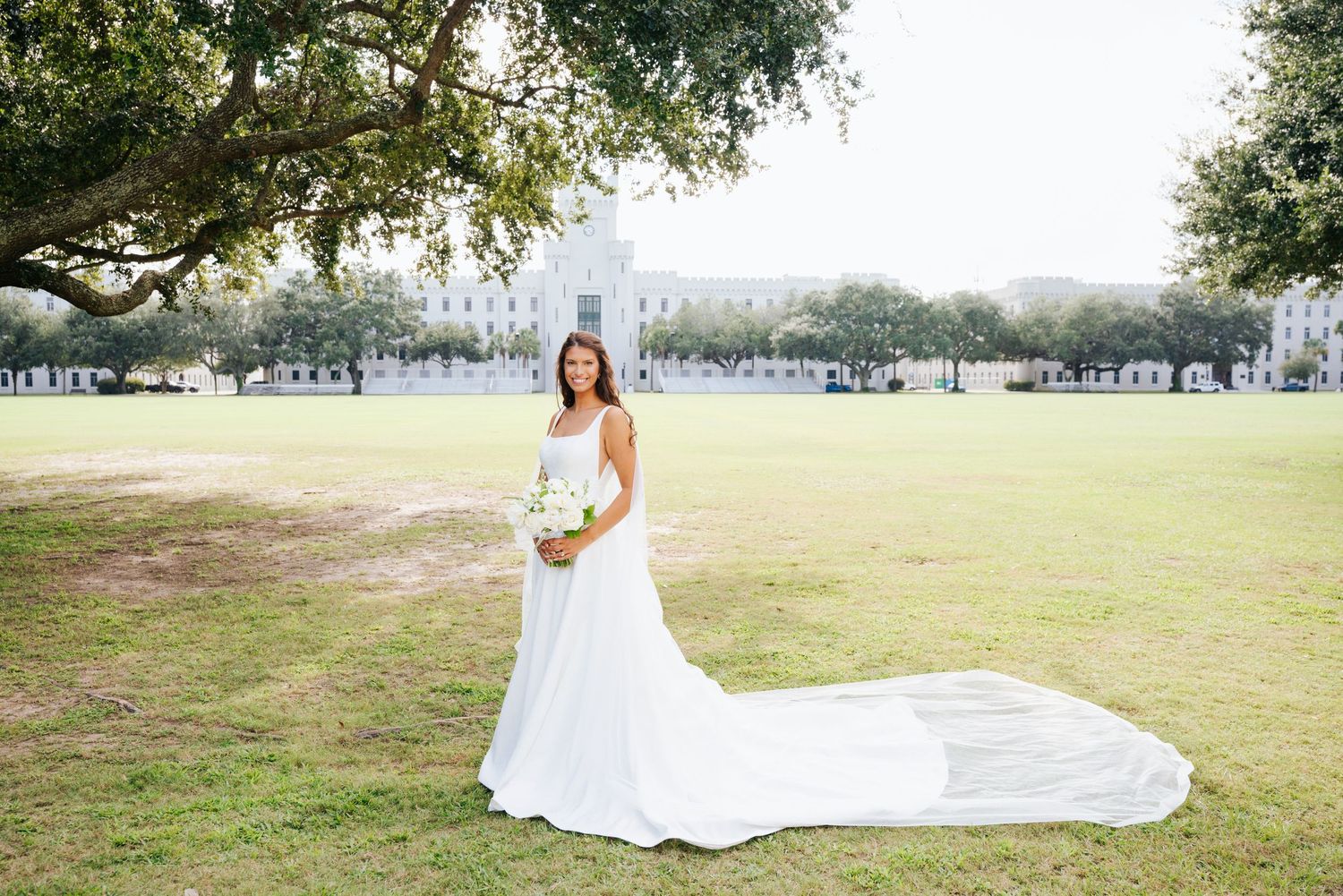 Long wedding dress train trailing across green grass with the Citadel barracks in the background.