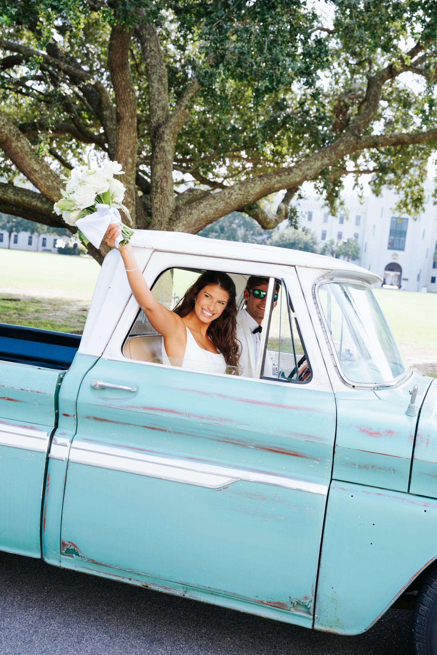 A bride poses playfully in a vintage mint green truck under oak trees.