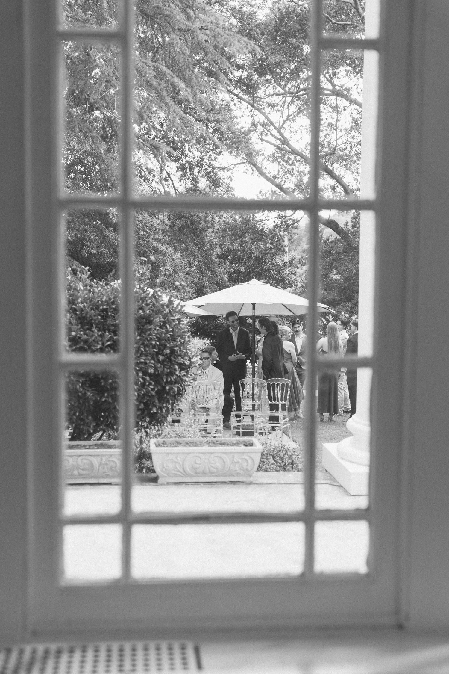 Black and white view through French doors of an outdoor wedding ceremony with guests under a parasol.