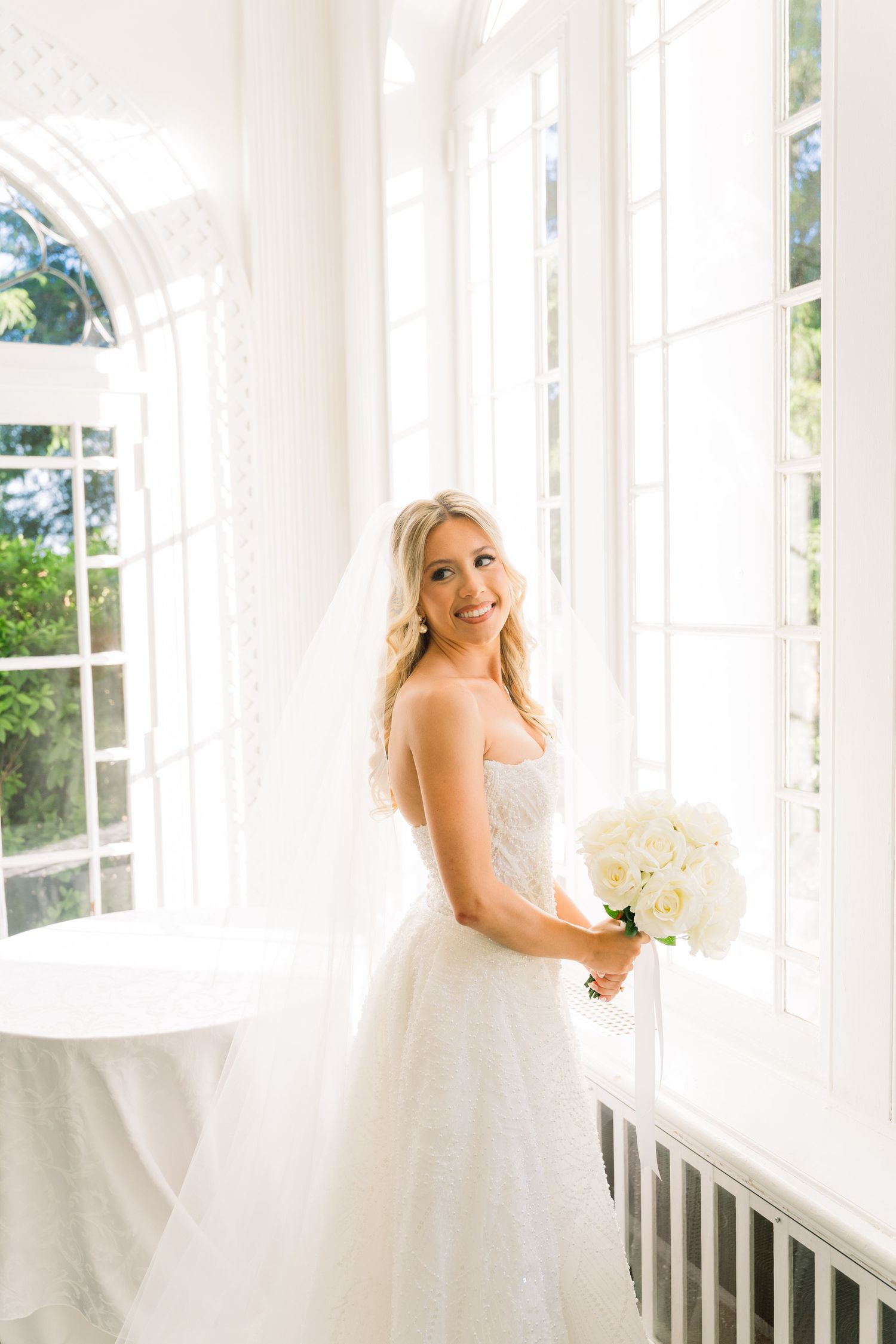 Smiling bride in strapless lace gown holding white bouquet stands by tall windows in bright elegant room.