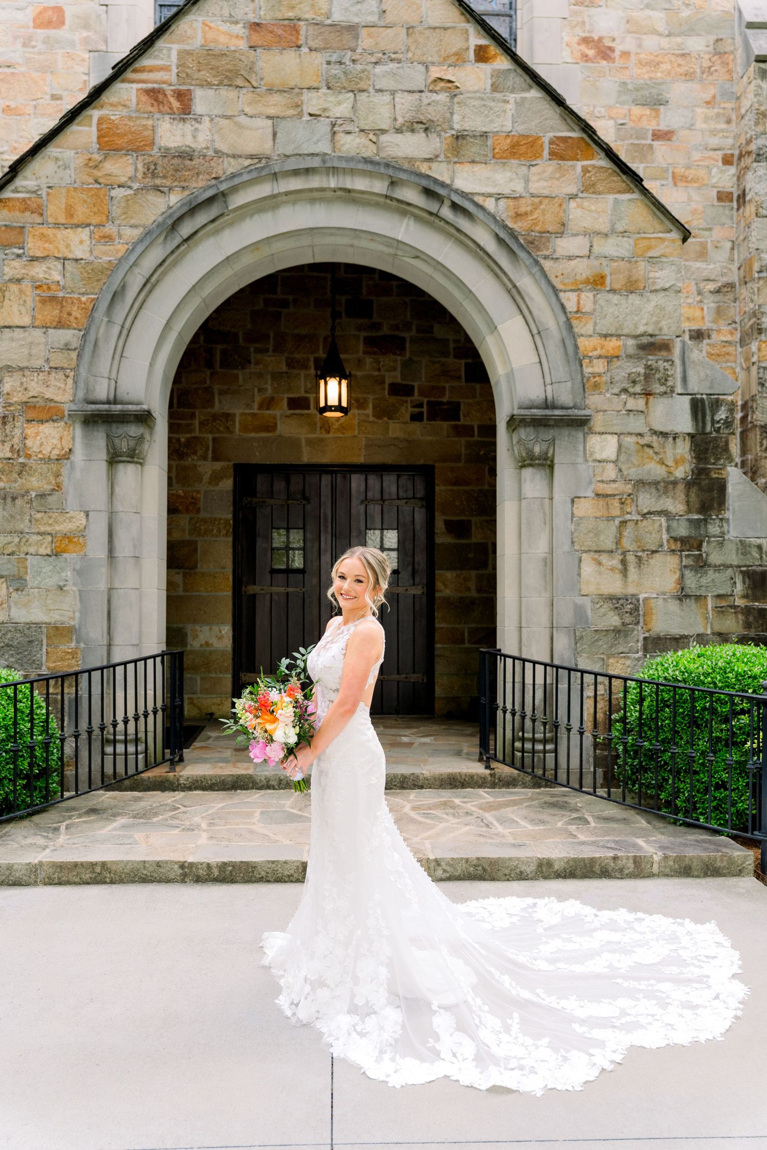 Bride in lace mermaid gown with cathedral train poses before stone church arched doorway holding colorful bouquet.