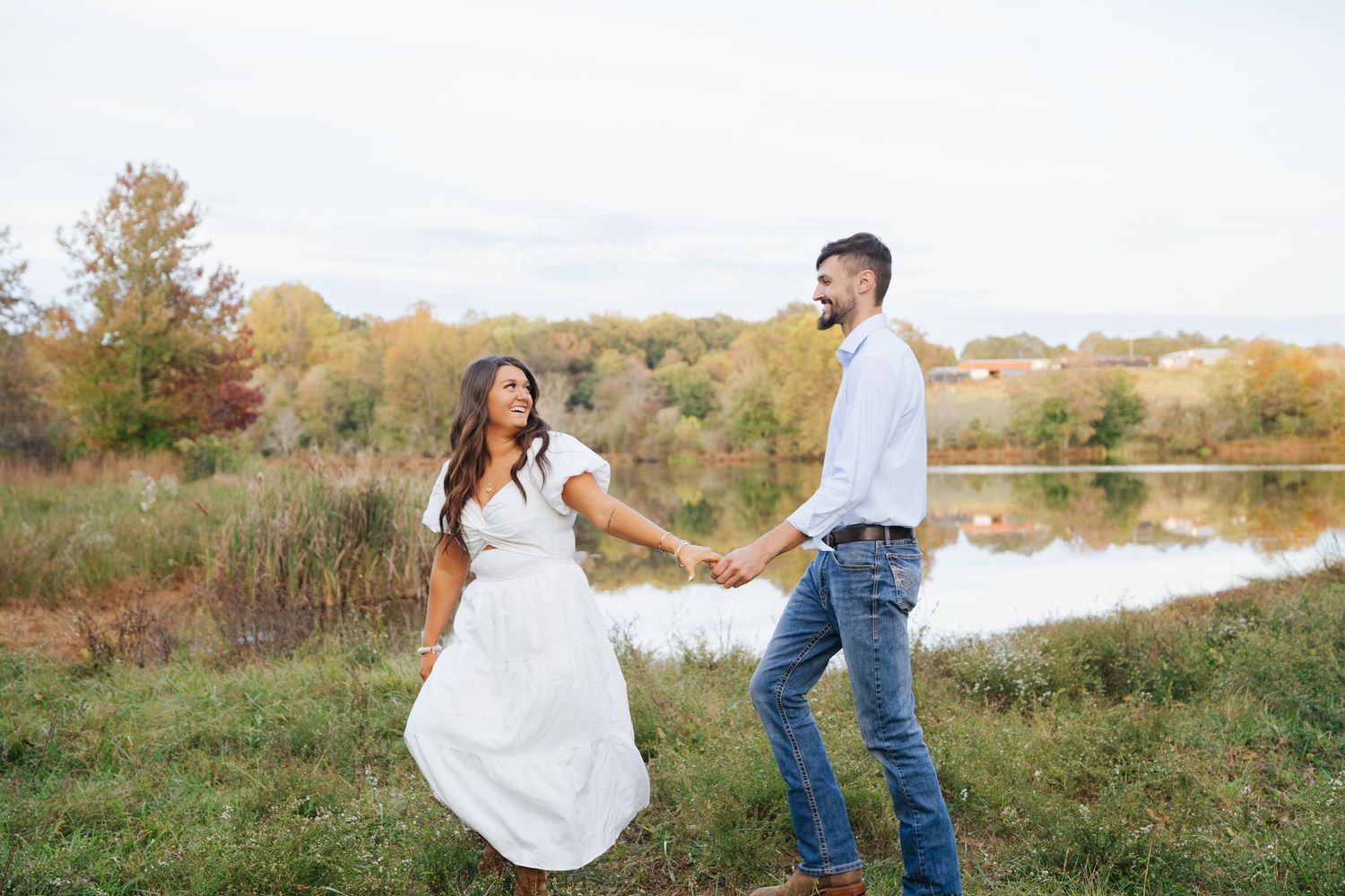 A couple holds hands in a romantic outdoor setting with autumn foliage and a lake in the background.