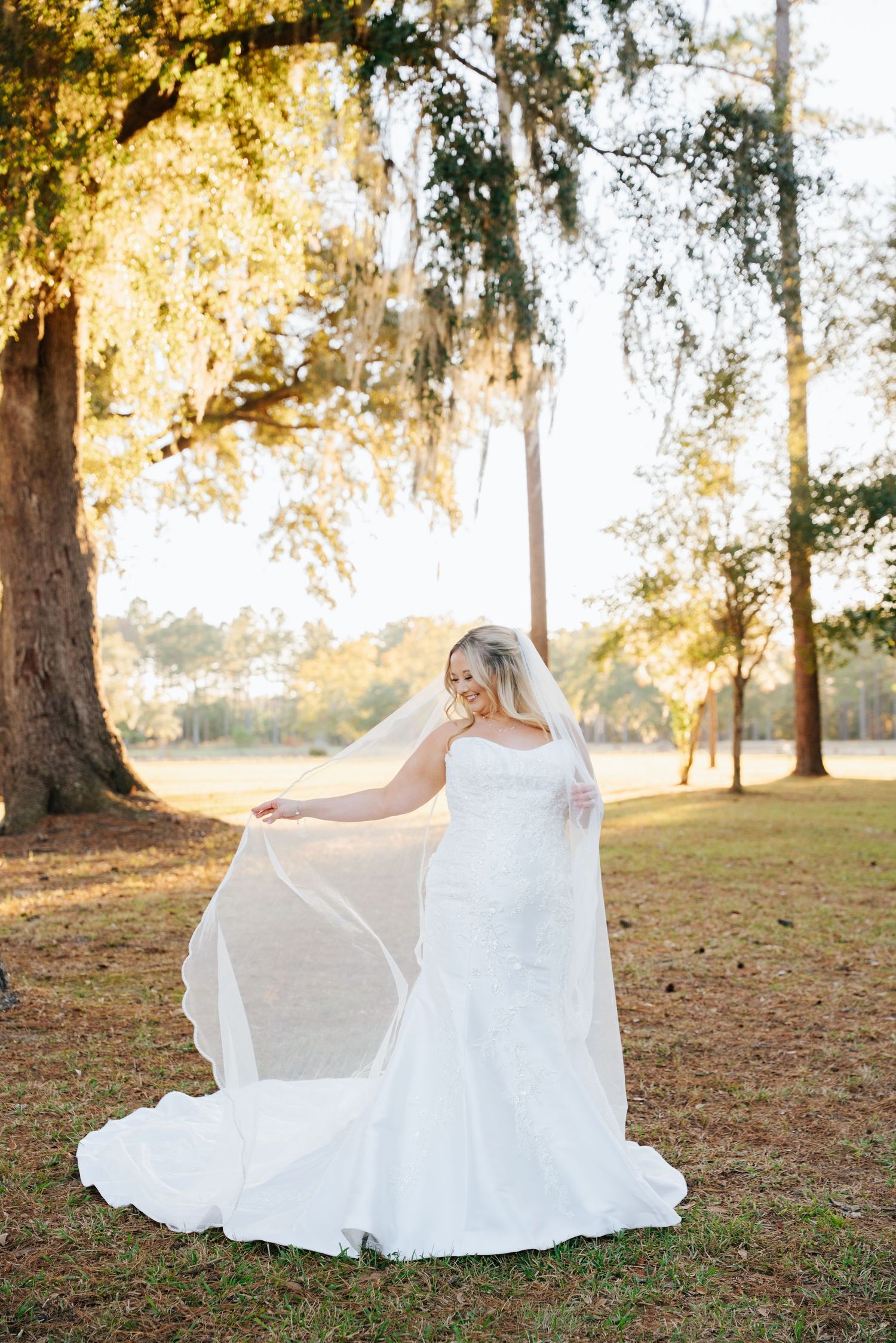 A flowing white wedding dress twirls gracefully in a sunlit park setting with tall trees in the background.