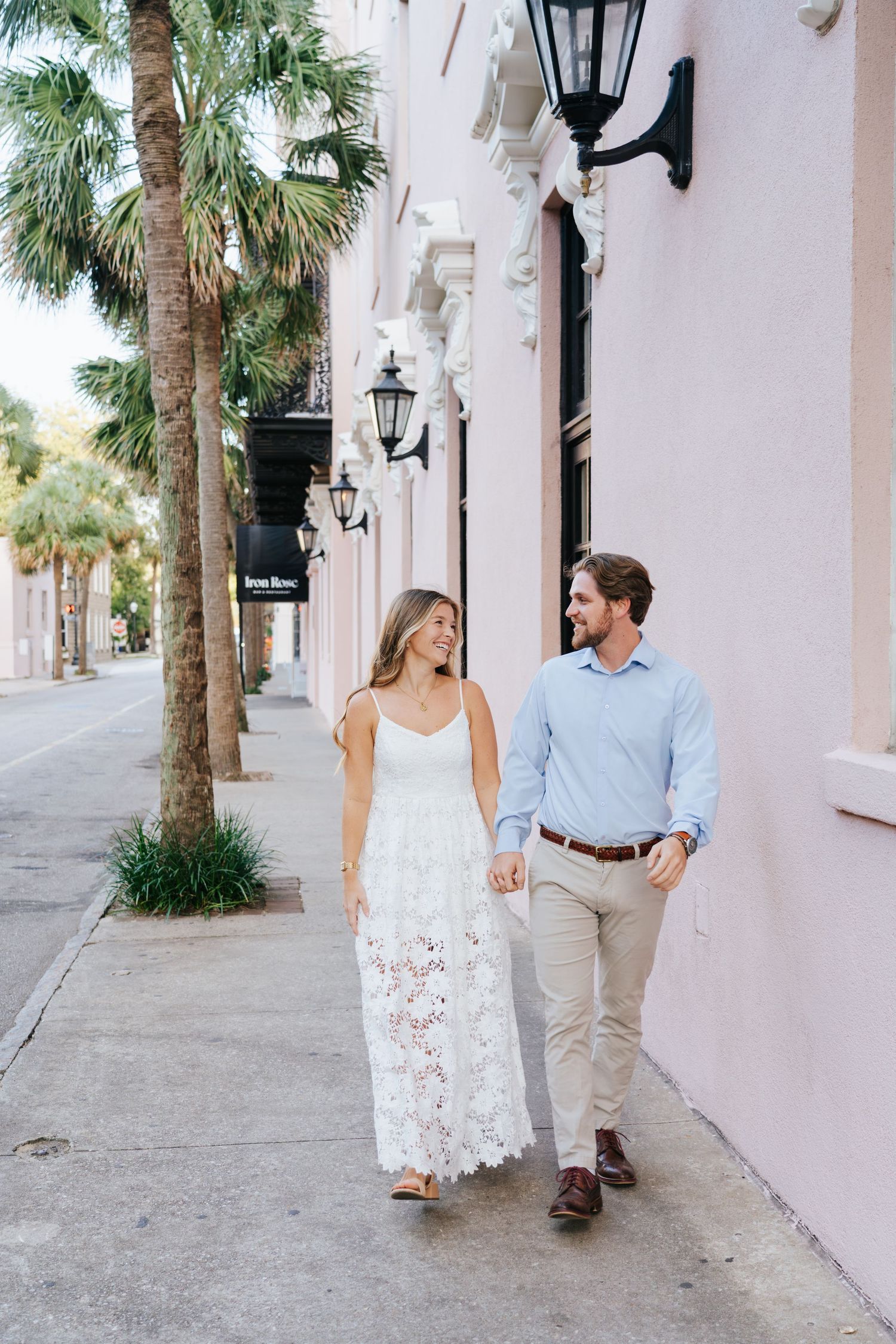 Couple strolls along pink-walled street lined with palm trees and vintage lanterns.