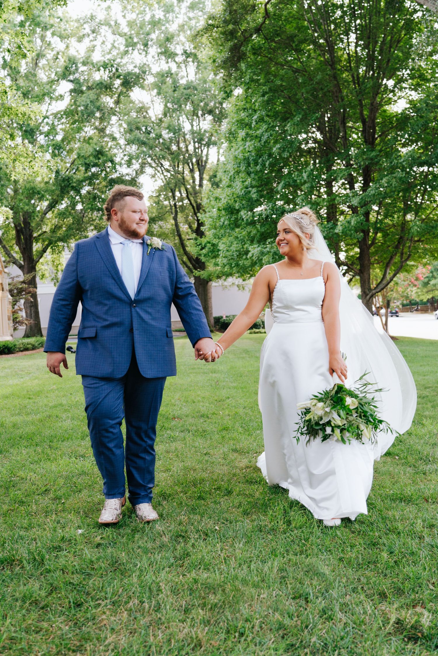 Playful wedding couple walking through a green lawn in blue suit and white dress.