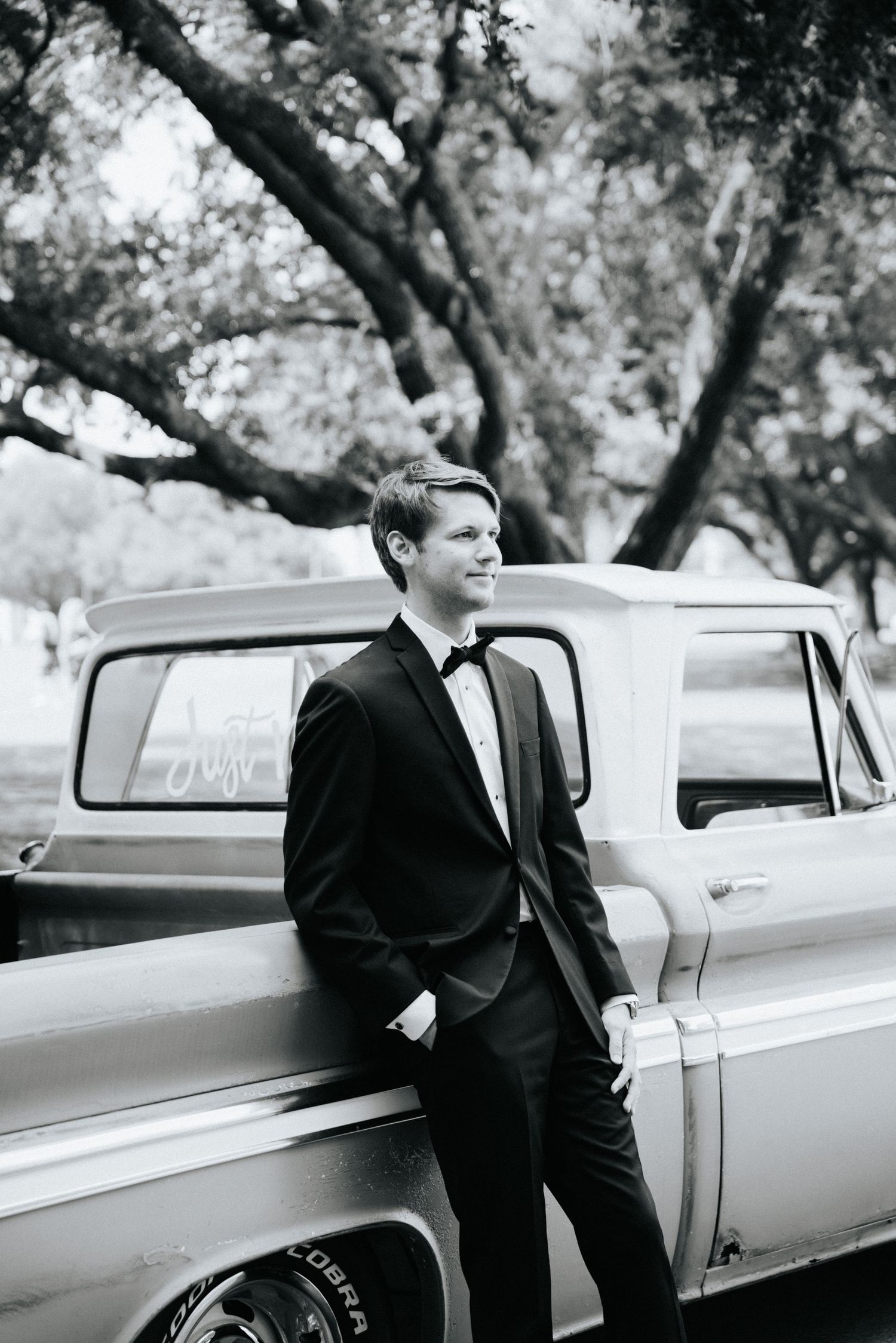 A groom in a black suit stands thoughtfully next to a classic truck in an outdoor setting.
