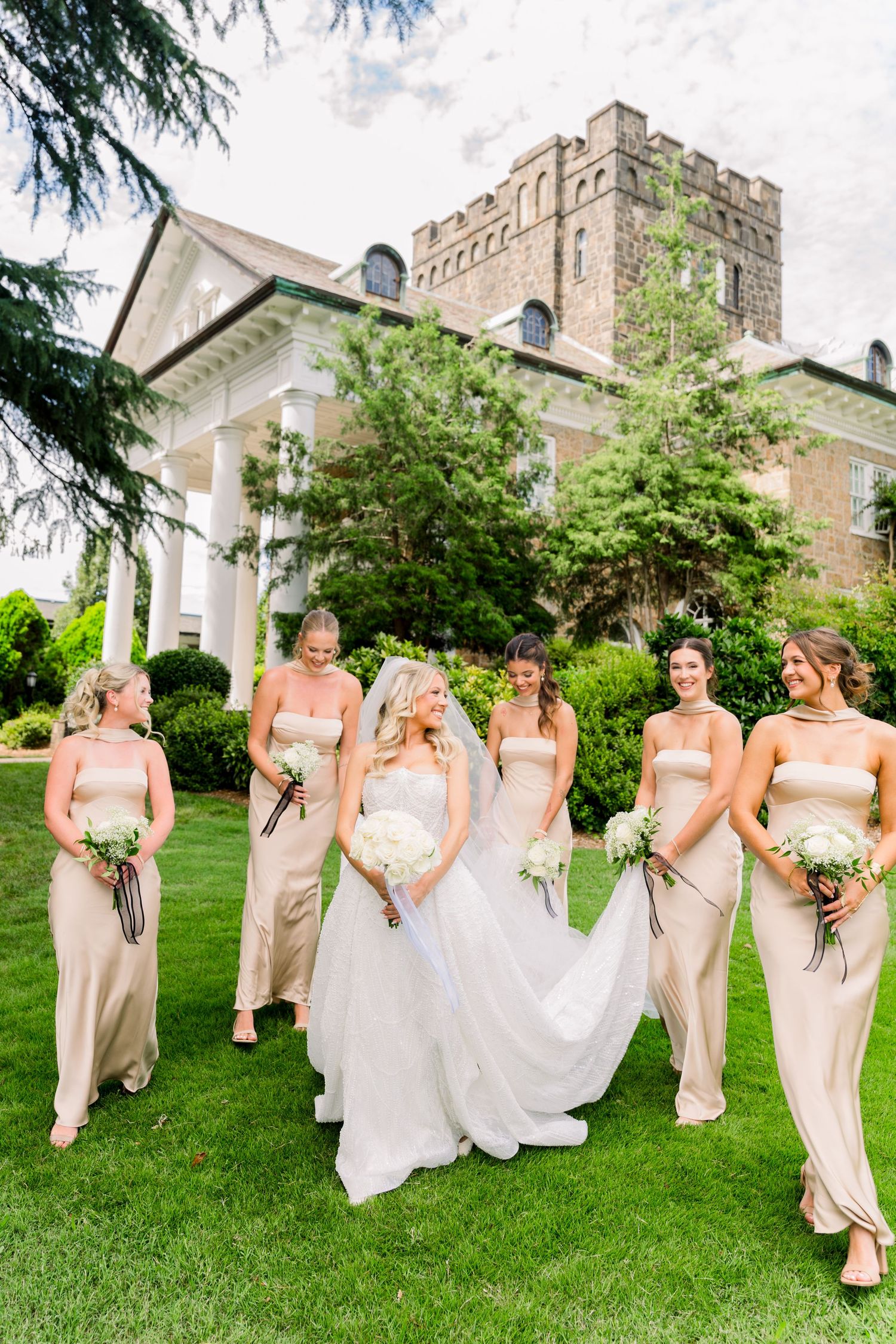 Bride in white gown walks with six bridesmaids in champagne dresses on green lawn before elegant estate.