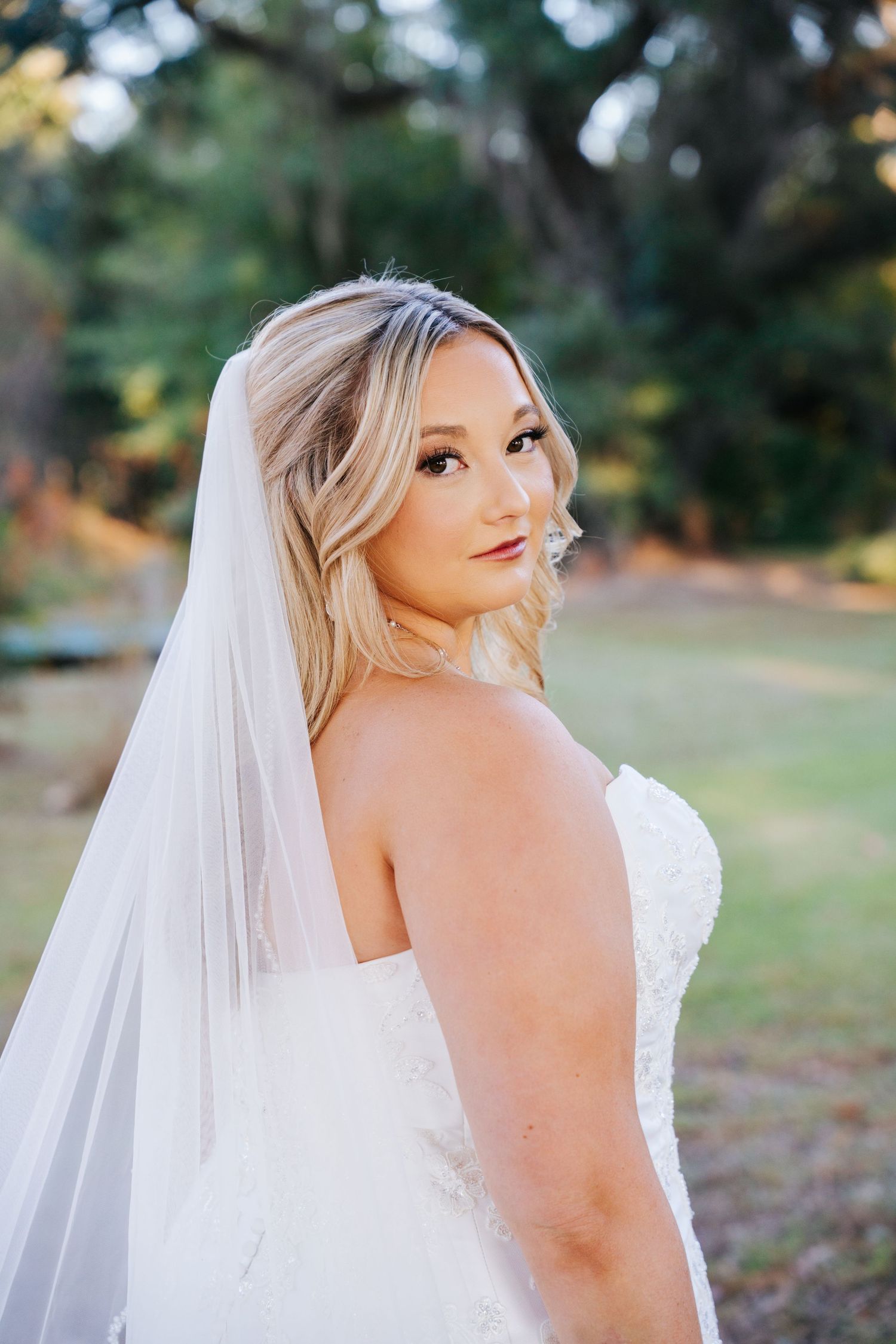 A bride in a strapless wedding dress and flowing veil looks over her shoulder in an outdoor setting.