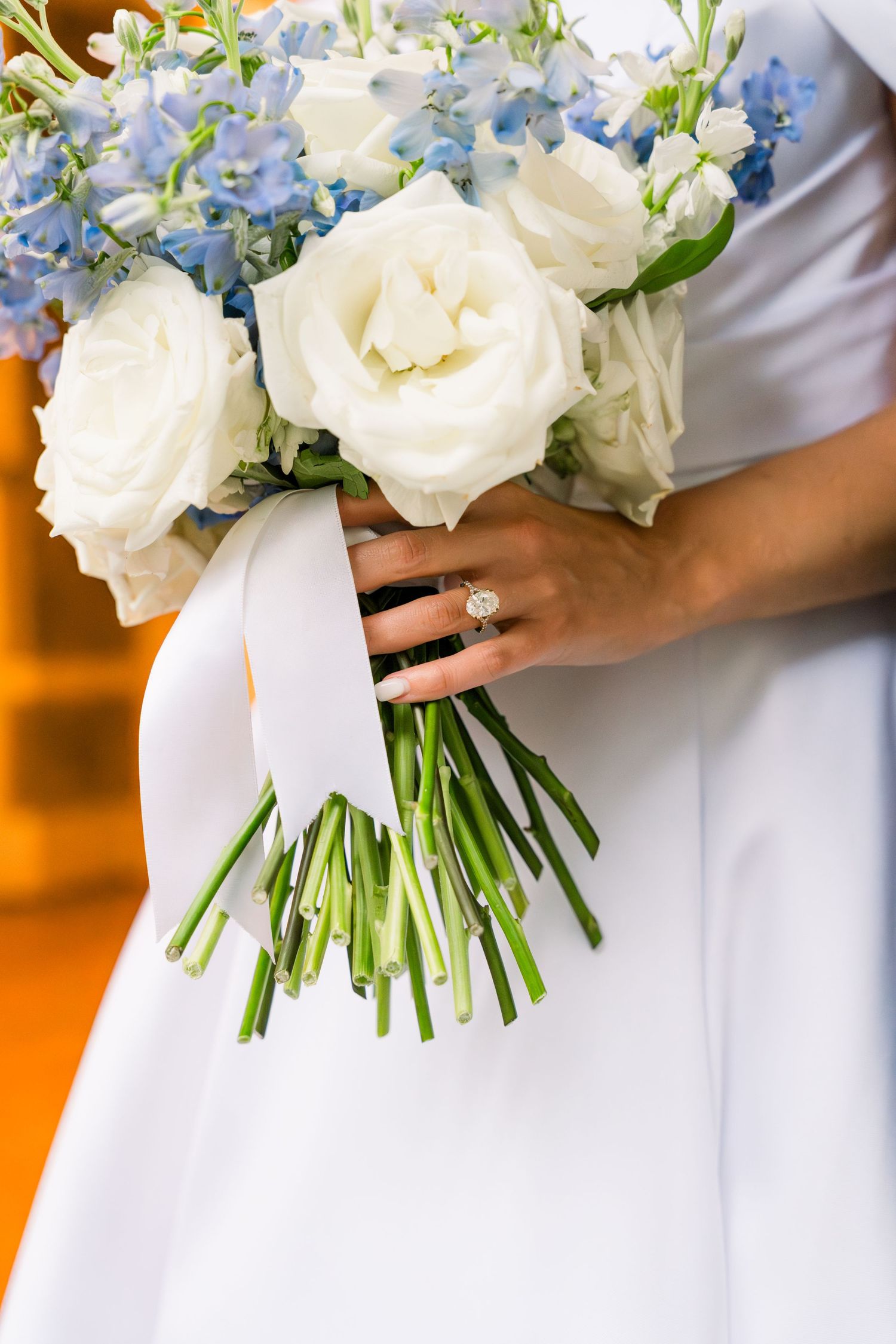 Bride holding a wedding bouquet of white roses and blue flowers tied with ribbon, wearing a diamond engagement ring.