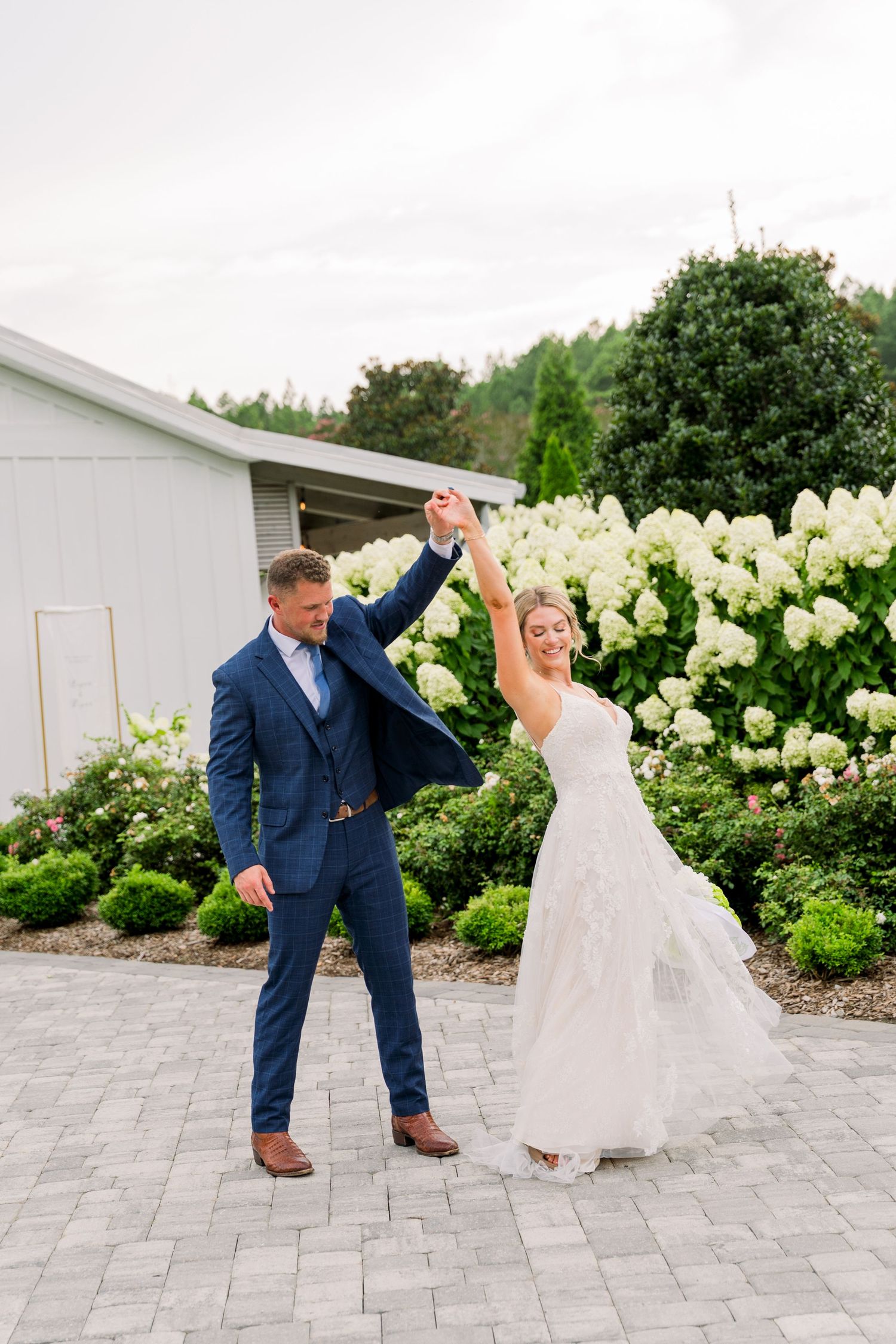 A joyful bride and groom dance outdoors on a paved patio surrounded by lush greenery and white hydrangeas.