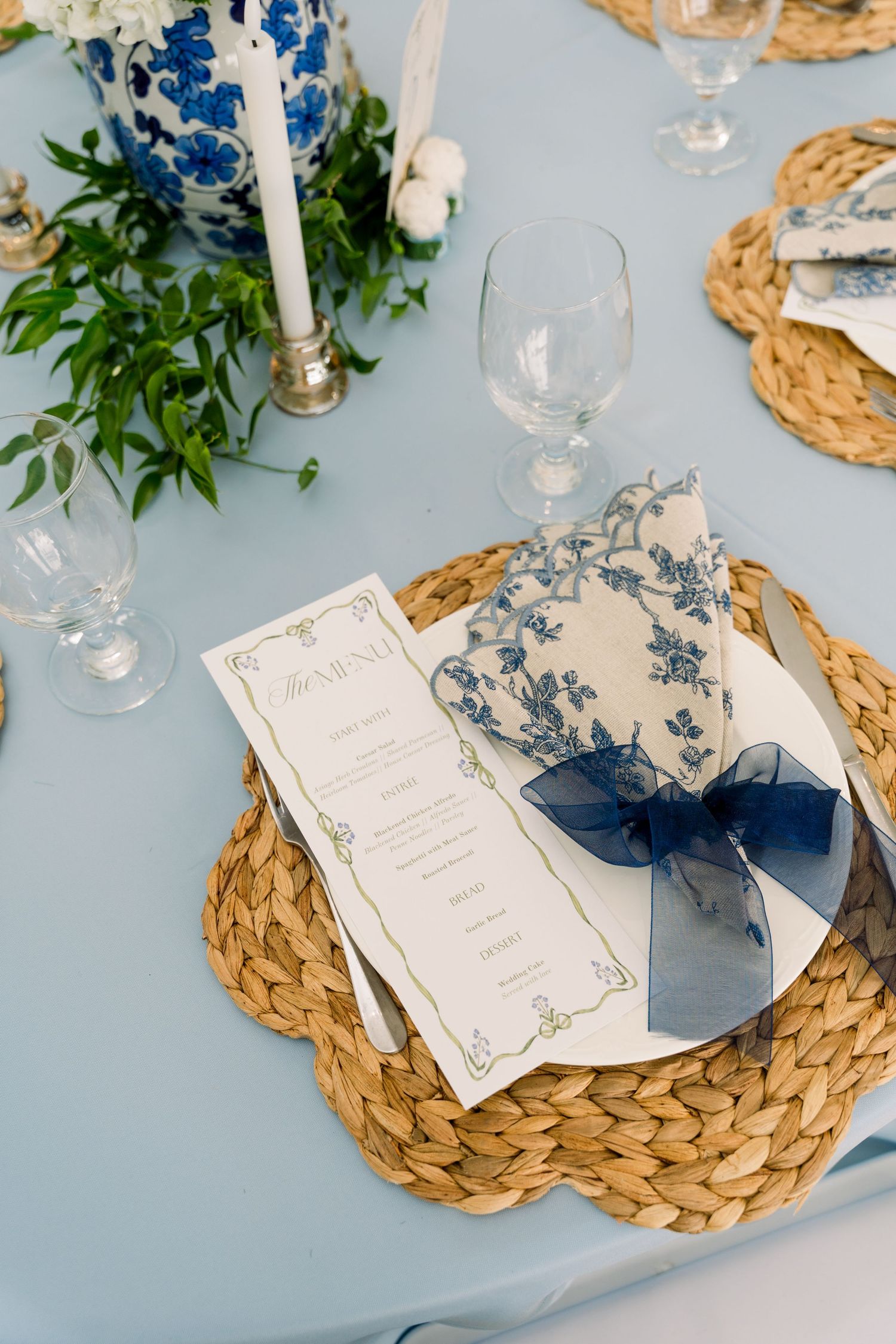 Elegant blue and white wedding table setting with woven placemat, floral napkin, navy bow, and menu card.