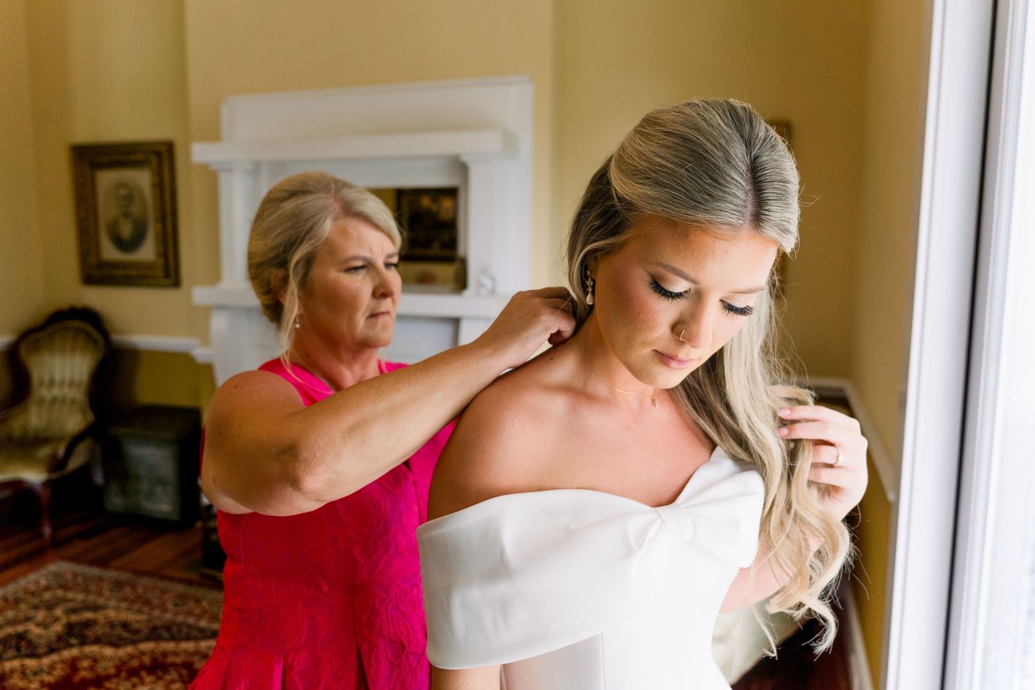 Mother helping bride with earrings while she adjusts her white wedding dress near a bright window.