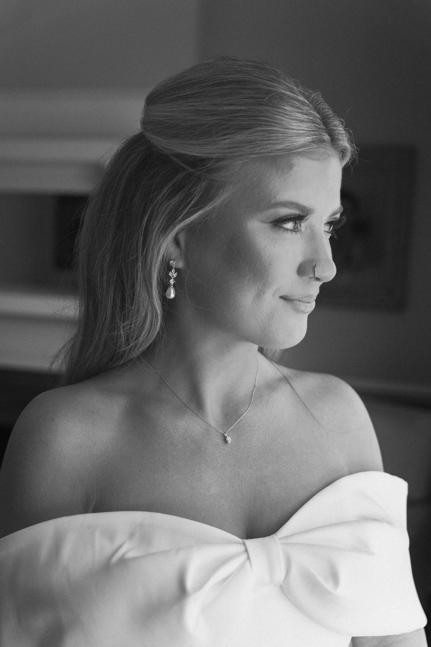 Black and white bridal portrait of a smiling blonde woman in an off-shoulder gown with pearl earrings and delicate necklace.