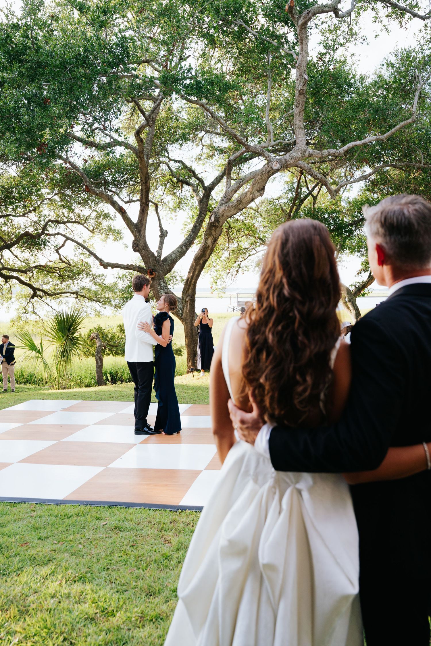 Bride and father sharing a heartfelt moment looking at groom and his mother dancing during their wedding reception under trees at sunset.