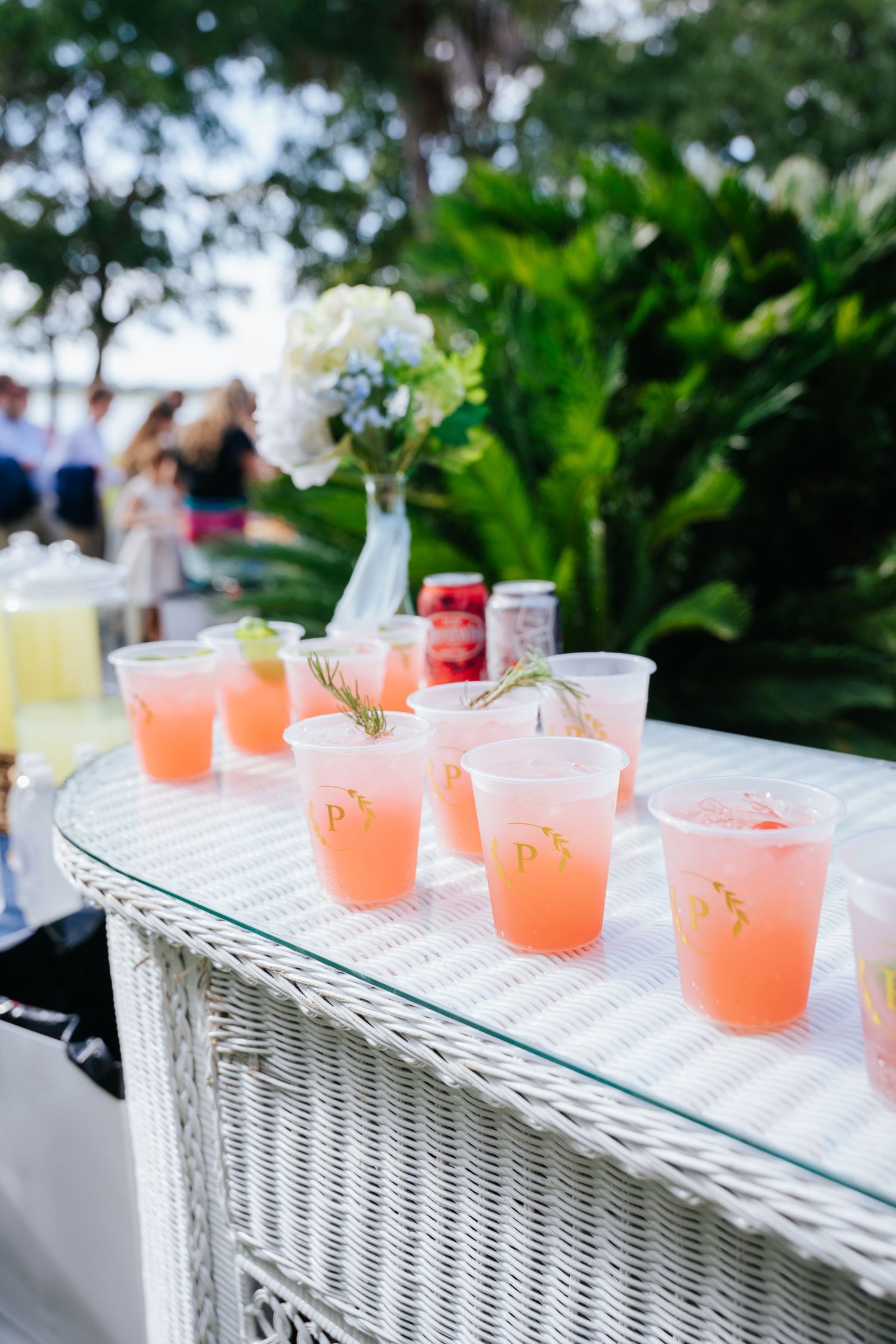 Pink mocktails in clear glasses arranged on a white wicker table at an outdoor reception.